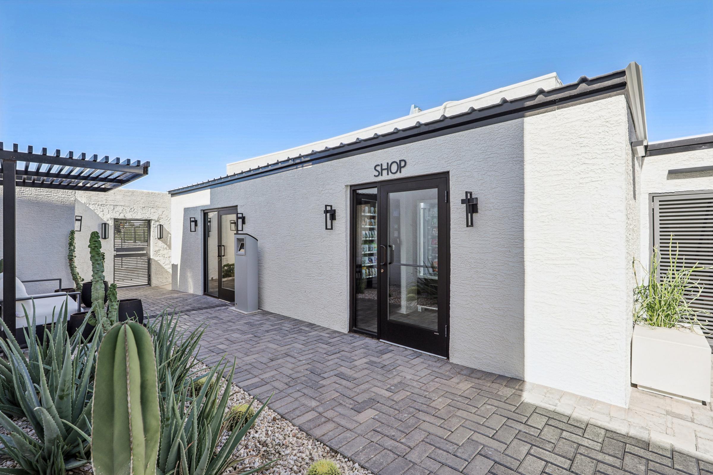 A modern shop building with a sign reading "SHOP" prominently displayed above the entrance. The exterior features a clean, white stucco finish with black accents, framed by potted plants and cacti. The area is well-lit with large windows, and the pathway leading to the entrance is paved.