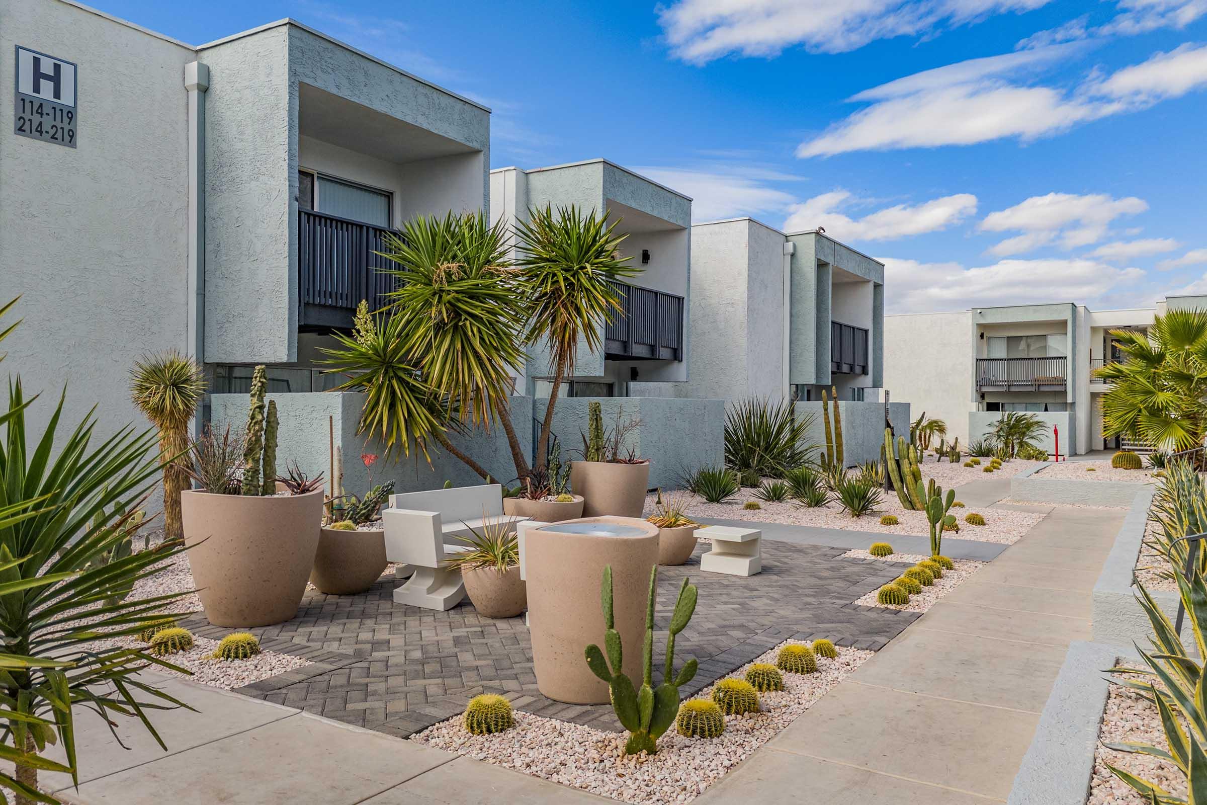 A landscaped courtyard featuring modern apartment buildings with balconies. The area includes a pathway lined with decorative stones, potted plants, cacti, and greenery. The sky is partly cloudy, creating a bright and inviting atmosphere.