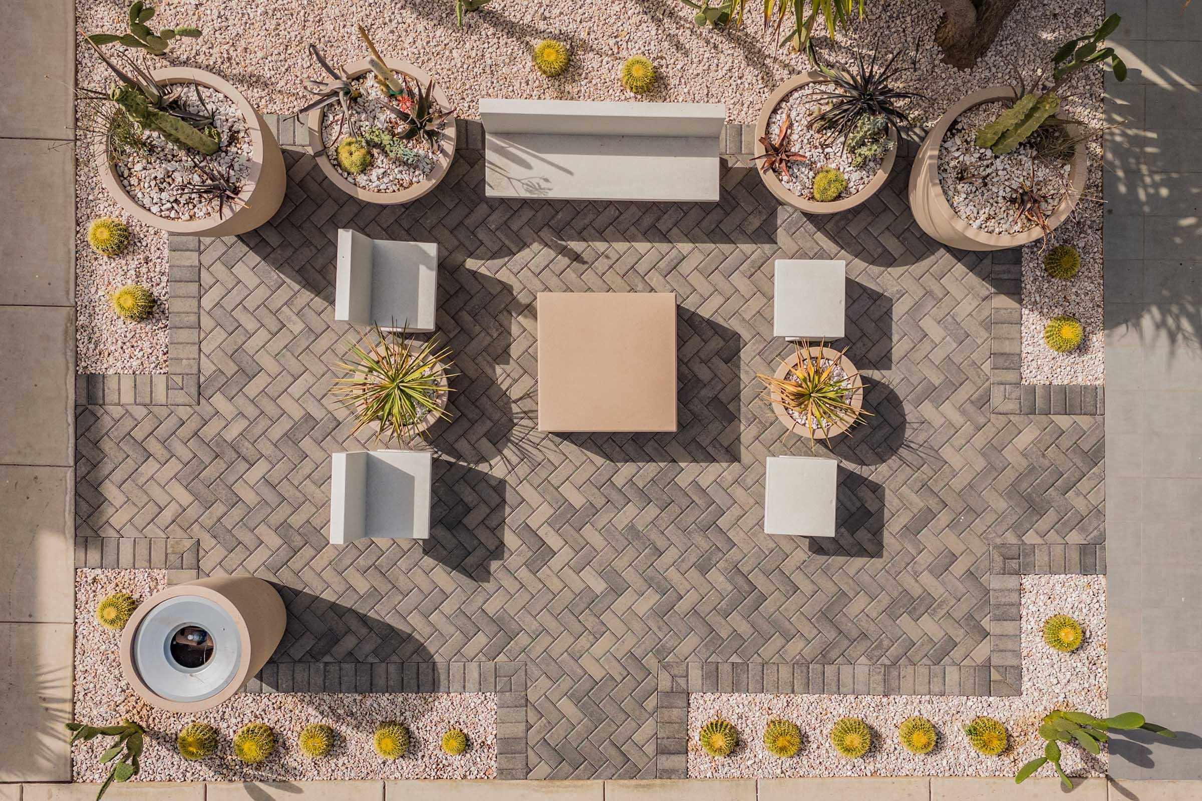 An aerial view of a modern outdoor patio with a herringbone-patterned stone floor. The area features circular planters filled with various cacti and succulents, yellow flowers, and a central square table surrounded by four light gray chairs. The setting is well-lit and designed for relaxation.