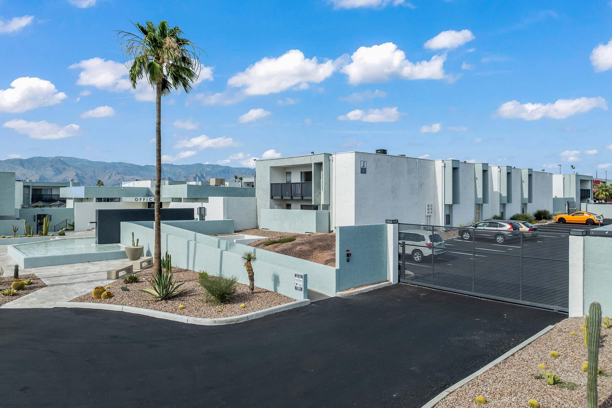 Modern apartment complex with light blue buildings, surrounded by landscaped areas and desert plants. A gated entrance is visible, along with a parking lot featuring several cars. In the background, mountains rise under a partly cloudy sky, and a tall palm tree adds a tropical touch.