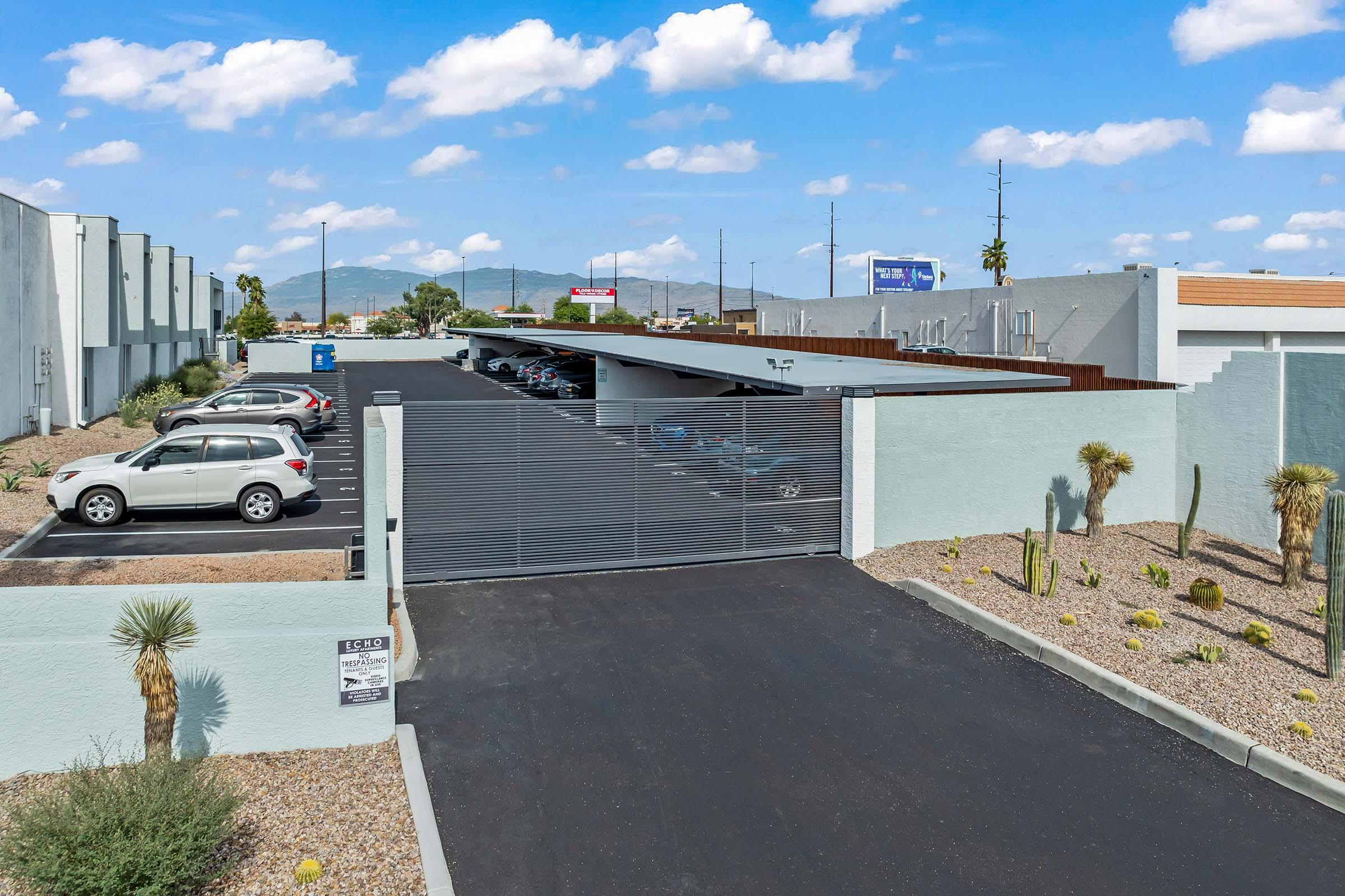 An outdoor parking lot featuring a gated entrance, with several parked cars under a shaded structure. Cacti and gravel landscaping border the area, while a clear blue sky with fluffy clouds is visible in the background. The setting is surrounded by buildings and distant mountains.