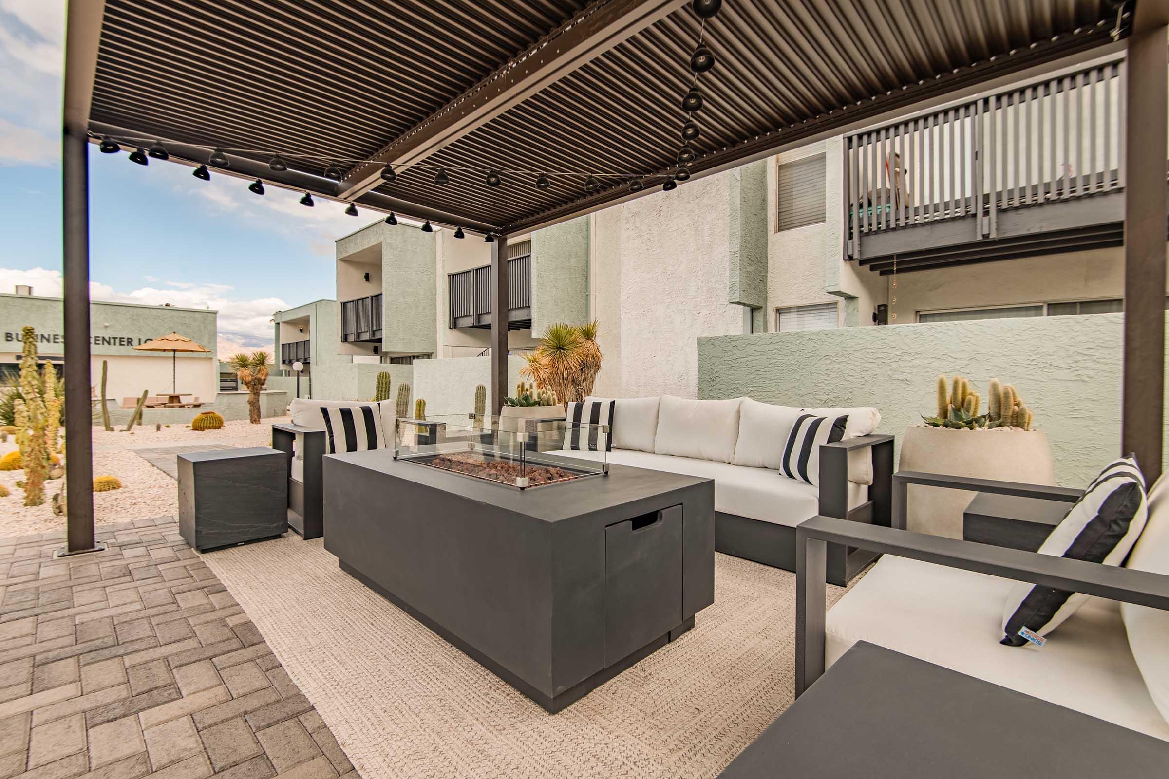 A modern outdoor seating area featuring a large, rectangular fire pit table surrounded by white cushioned sofas with black accent pillows. The space is shaded by a pergola with recessed lighting, set against a backdrop of contemporary buildings and desert landscaping.