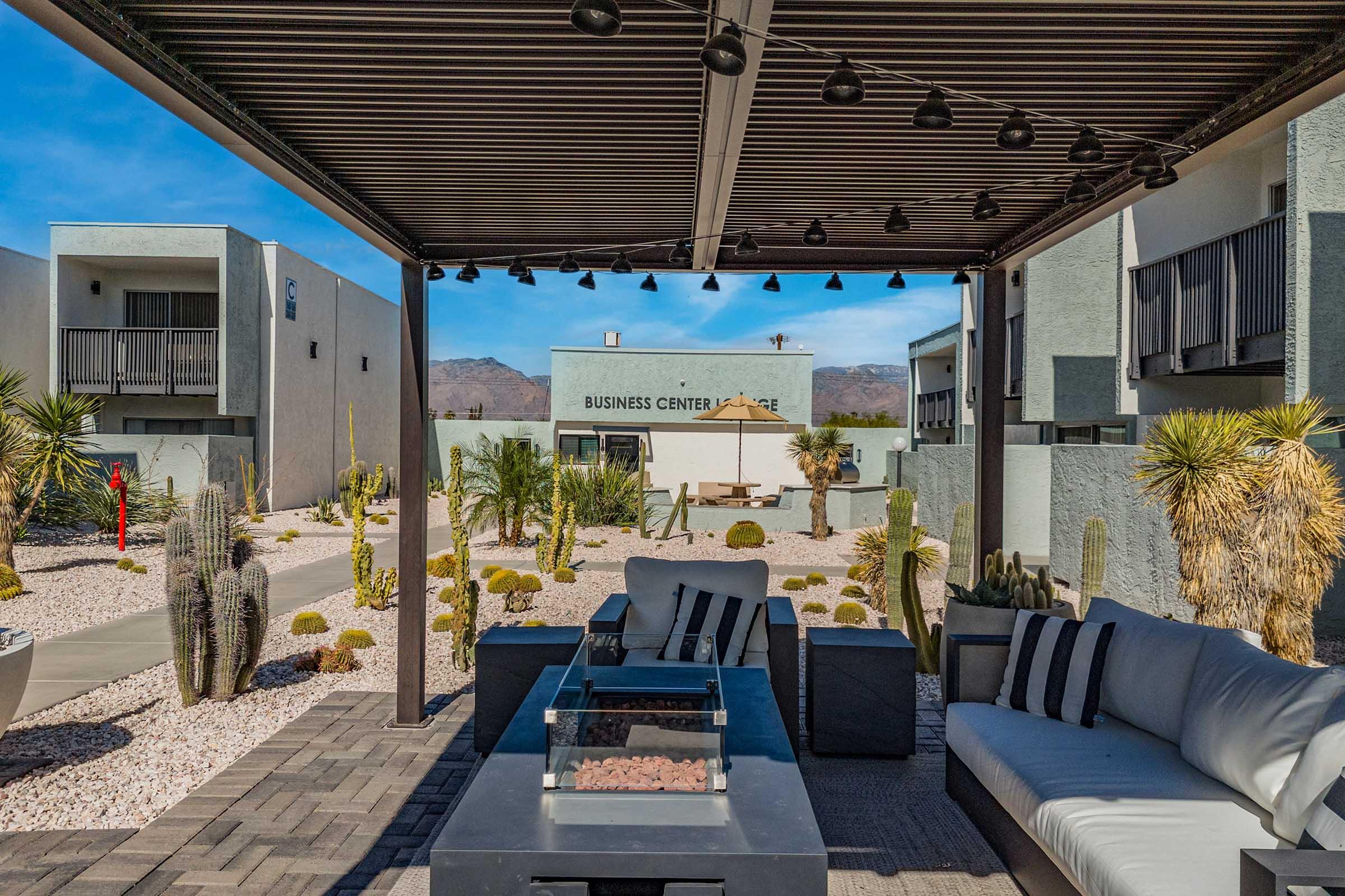 A modern outdoor seating area with a sectional sofa and coffee table, surrounded by landscaped desert plants and gravel. In the background, a building labeled "Business Center" is visible, with mountains in the distance under a clear blue sky. The space is shaded by a pergola with hanging lights.