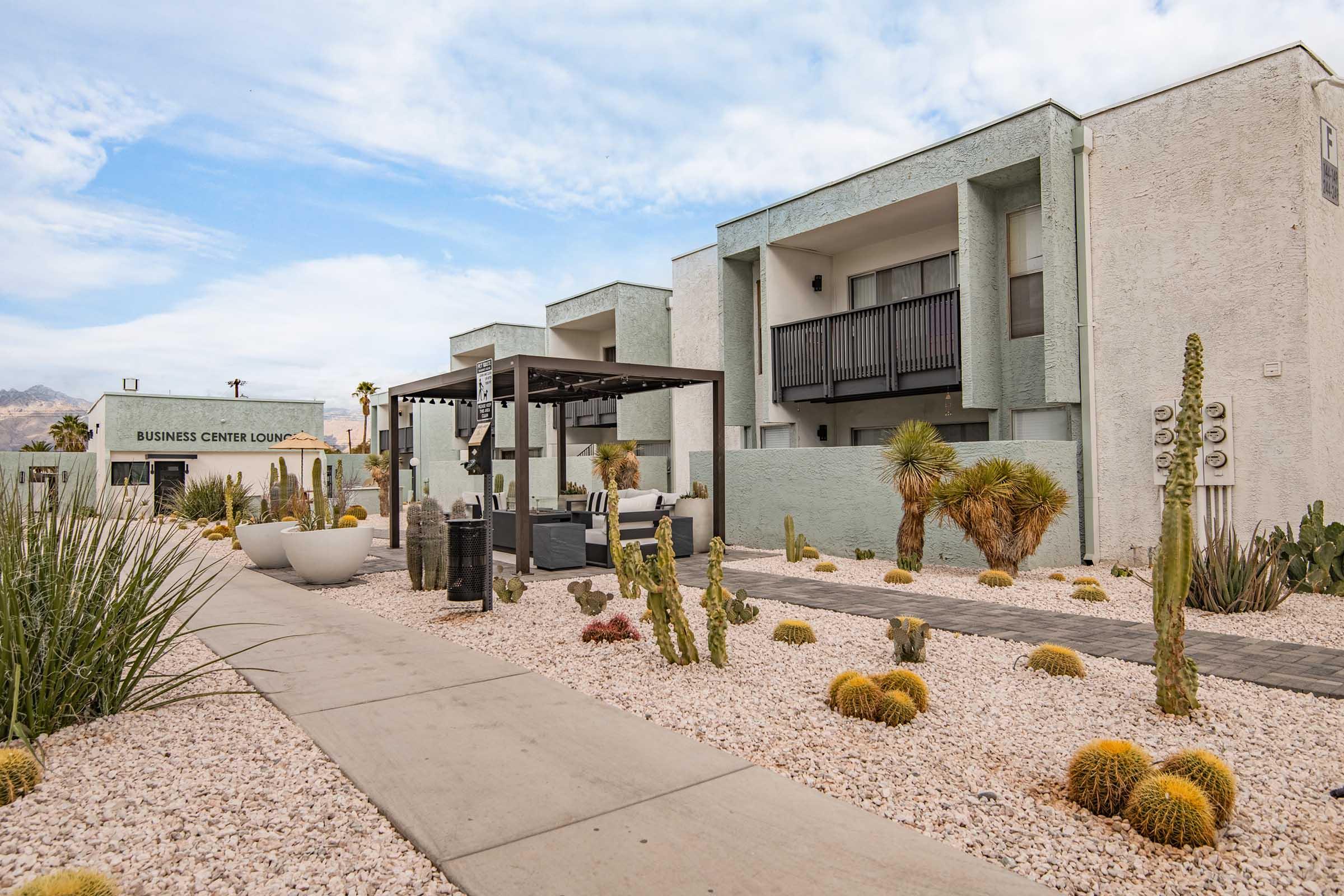 A modern, light-colored building with balconies is set against a clear sky, surrounded by desert landscaping featuring various cacti and decorative rocks. In the foreground, there's a pathway leading to a seating area under a pergola. The business center is visible to the left.