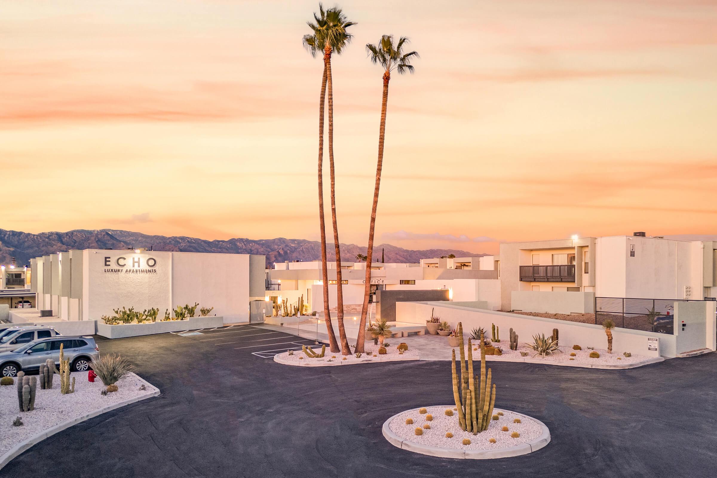 A modern resort named "ECHO" featuring minimalist architecture surrounded by desert landscaping. Tall palm trees and a circular driveway are prominent, with mountains in the background under a colorful sunset sky. Cacti and gravel add to the desert ambiance.