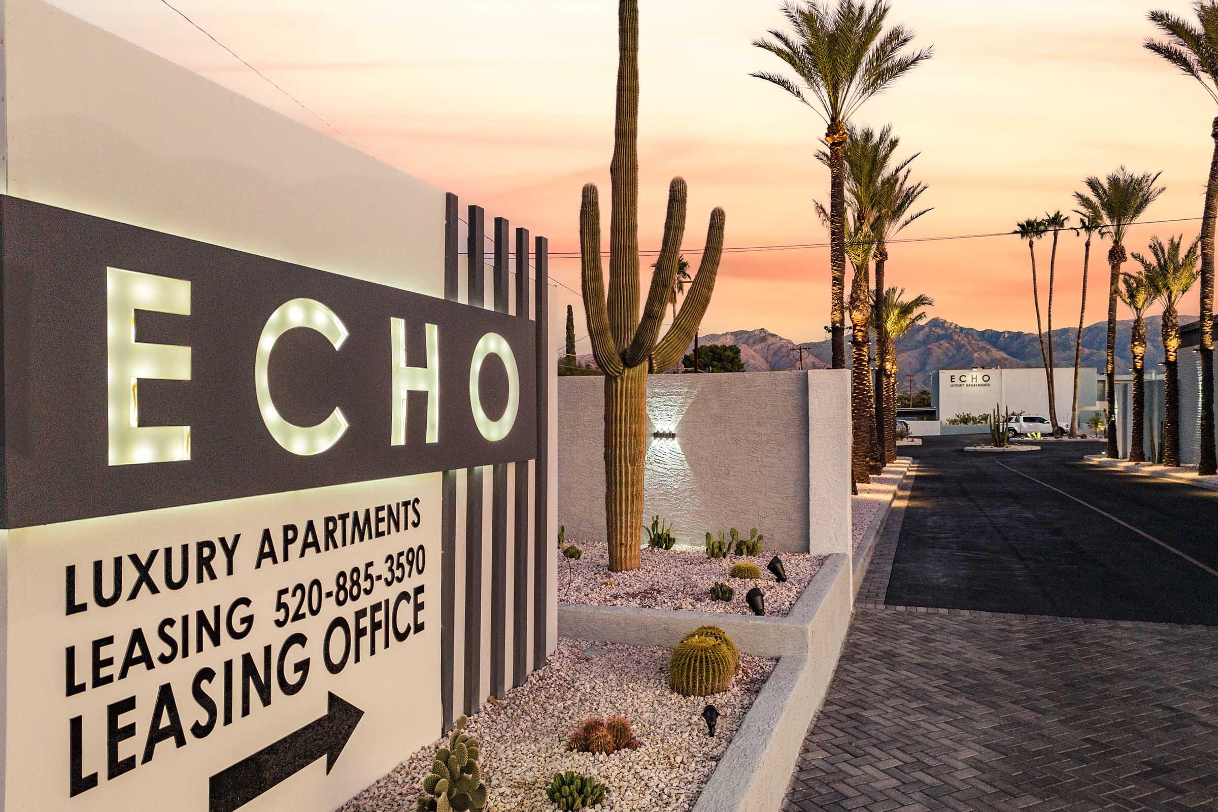 Sign for Echo Luxury Apartments featuring large illuminated letters. Desert landscaping with cacti and gravel. The leasing office contact number is visible. The background shows mountains and a sunset sky, indicating a warm, inviting atmosphere.