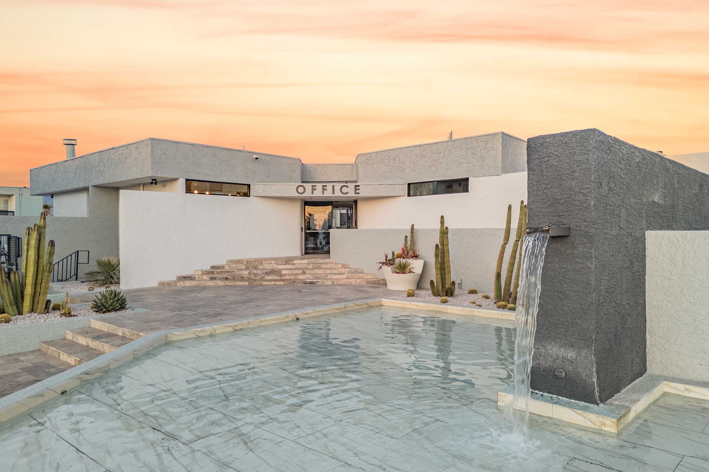 A modern office building with a minimalist design, featuring a staircase leading to the entrance labeled "OFFICE." In front, there's a tranquil water feature with a small waterfall, surrounded by potted plants and cacti, all set against a vibrant sunset sky.