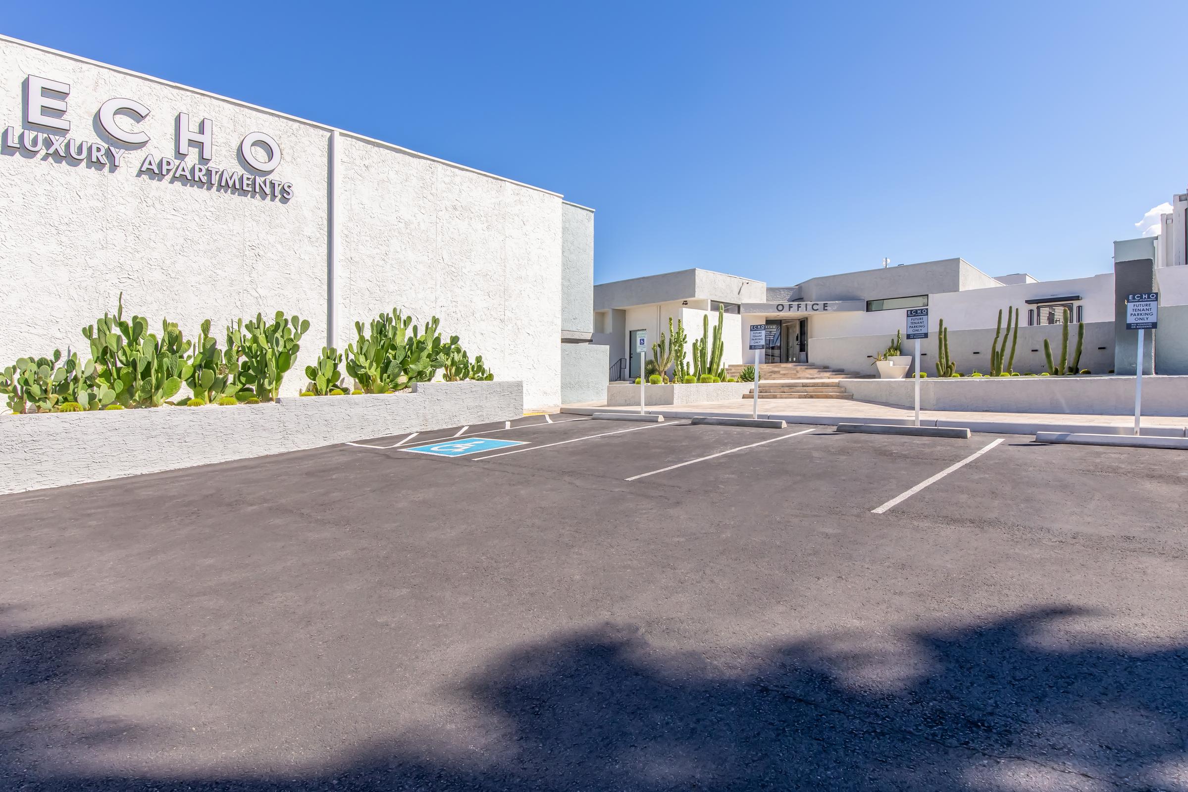 A view of the entrance to Echo Luxury Apartments, featuring a well-maintained exterior with cacti along the side. The parking lot is visible in the foreground with designated spaces and a clear blue sky overhead. An office area is seen in the background, adding to the welcoming atmosphere of the complex.