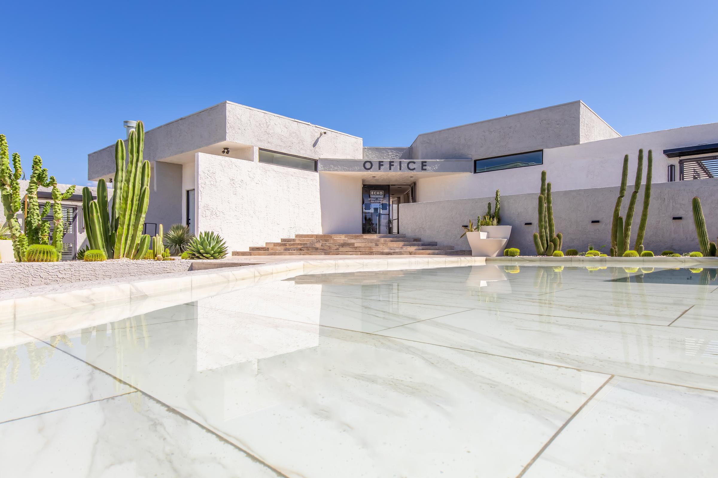 Modern office building with a minimalist design, featuring large glass windows and a flat roof. The entrance has a set of steps leading up to the word "OFFICE" displayed prominently. Surrounding the building are various cacti and succulents, with a reflecting pool in the foreground under a clear blue sky.