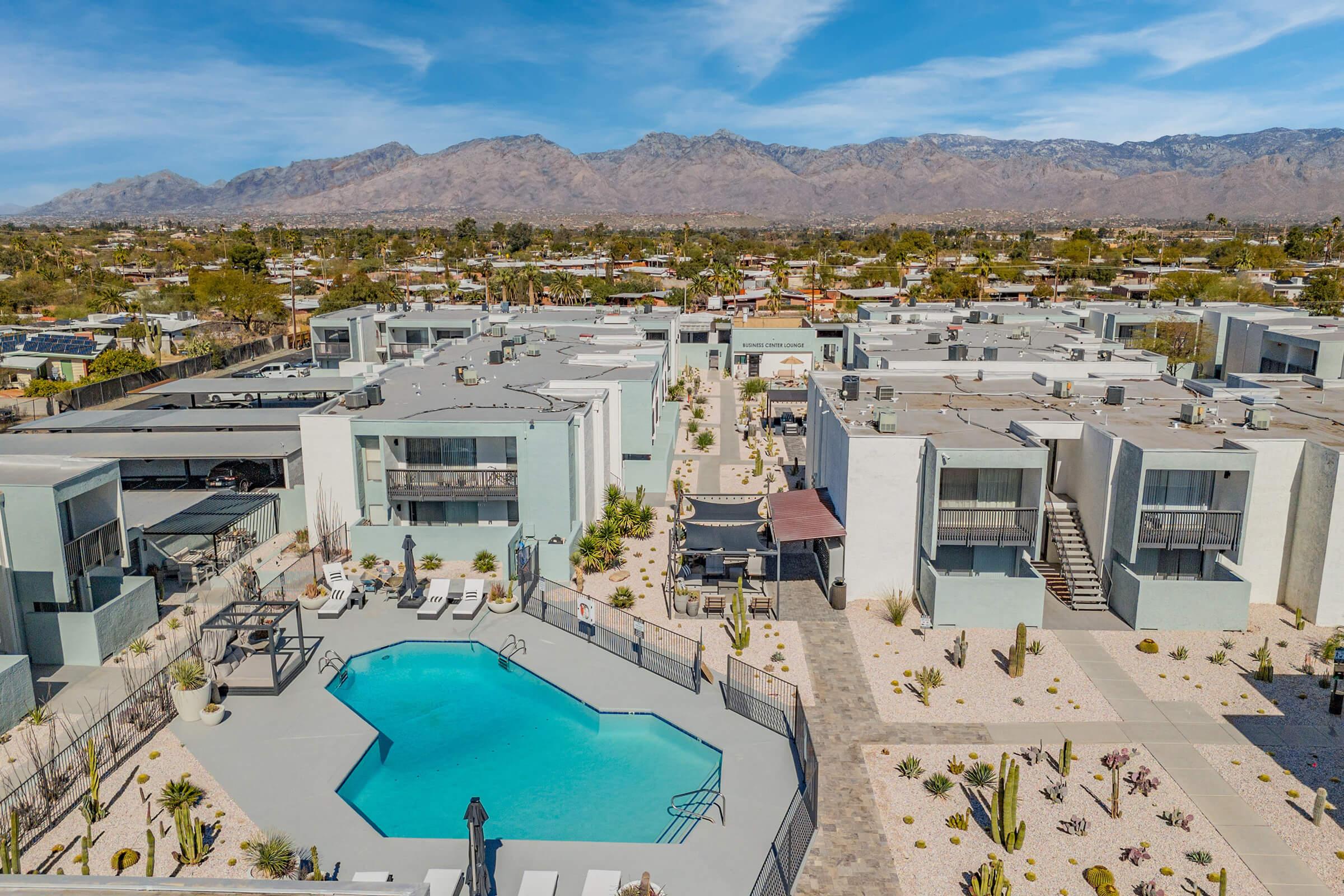 Aerial view of a modern apartment complex featuring a turquoise swimming pool surrounded by lounge chairs. The area is landscaped with desert plants, and distant mountains provide a scenic backdrop under a clear blue sky.