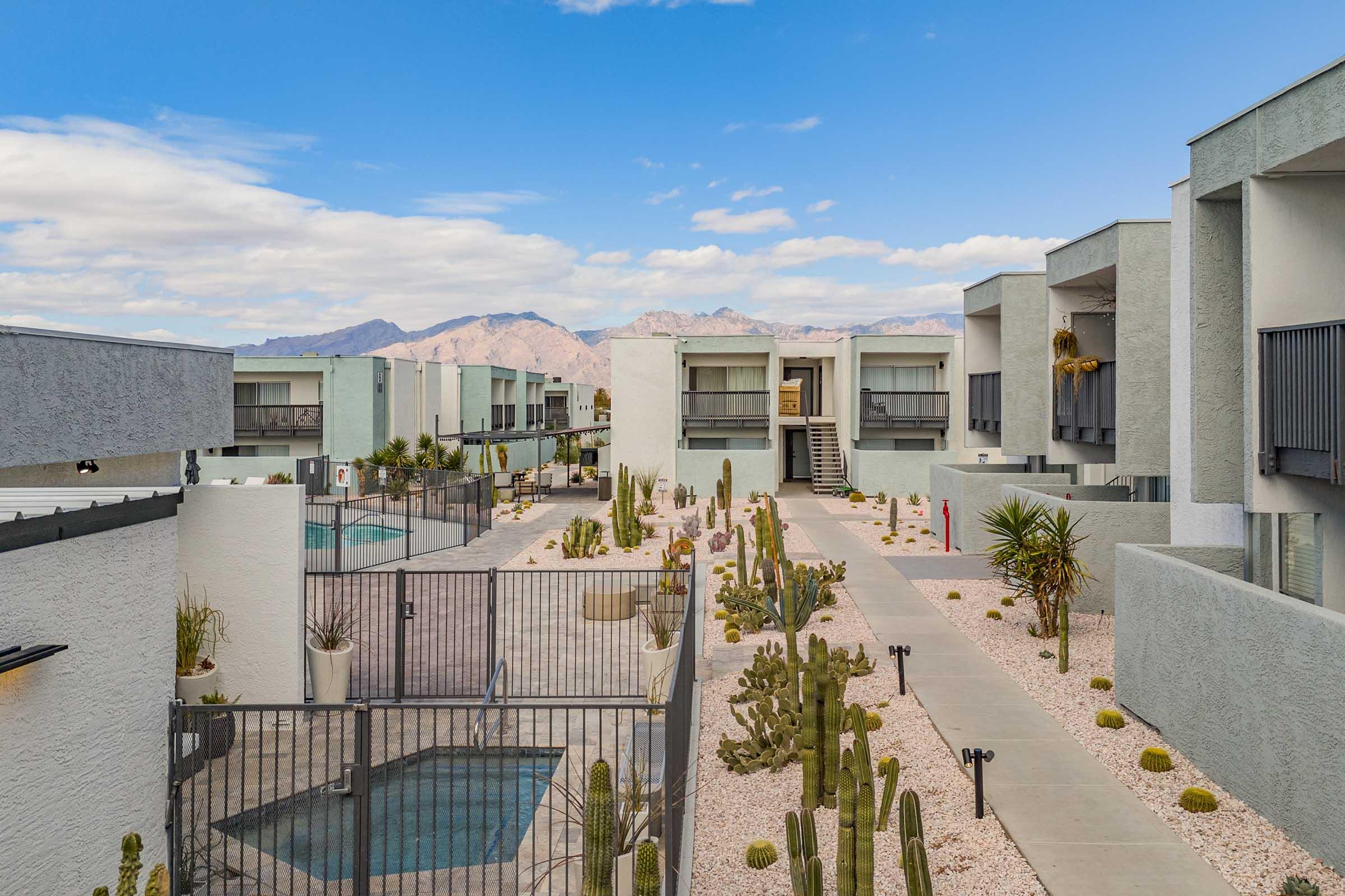 A view of a modern apartment complex featuring multiple buildings with outdoor pathways. The landscape includes desert plants and cacti, with a pool area visible behind a fence. The backdrop showcases mountains under a partly cloudy sky, creating a serene atmosphere.