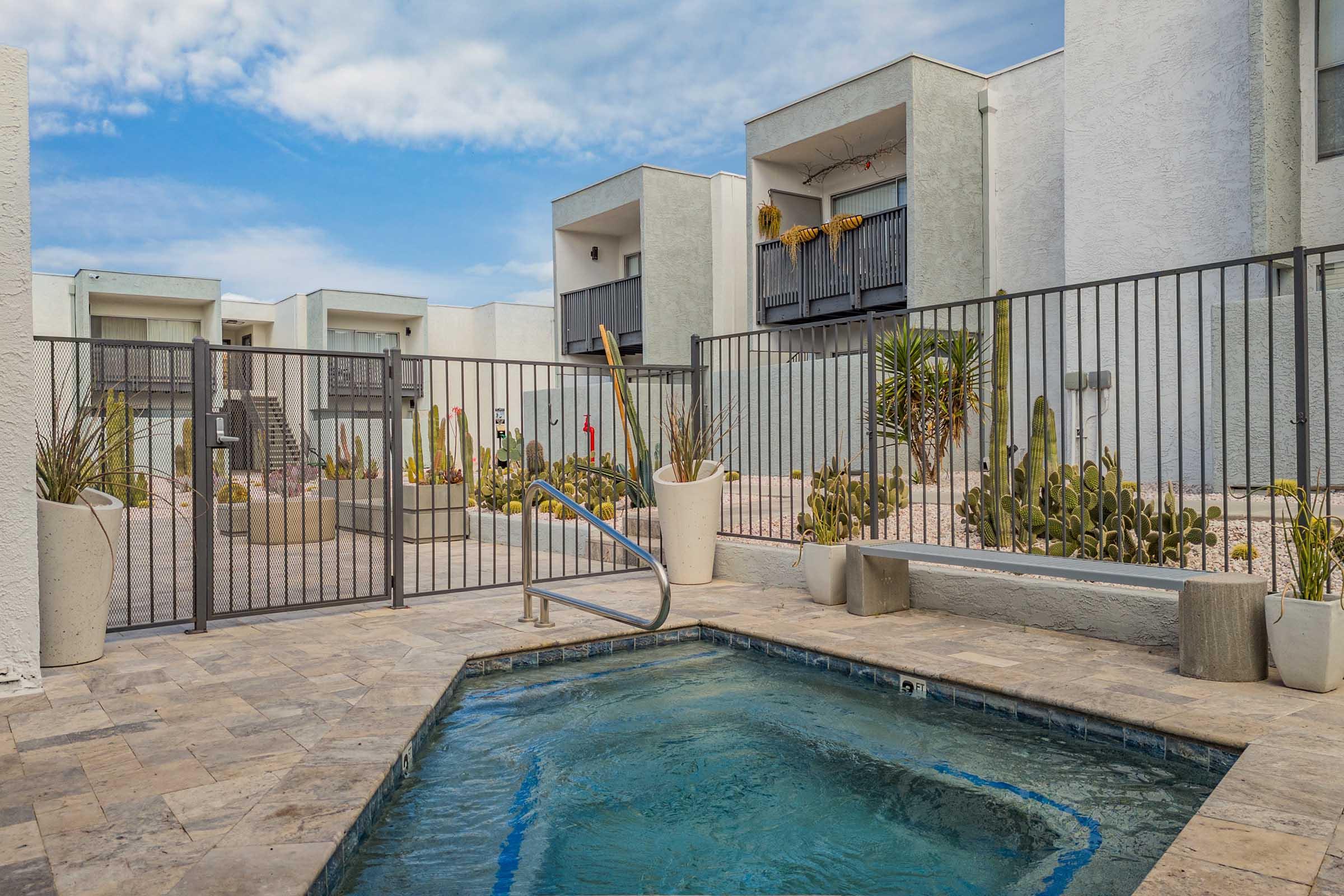 A gated pool area featuring a small swimming pool, surrounded by desert landscaping with cacti and stone pathways. In the background, modern apartment buildings with balconies are visible under a partly cloudy blue sky.