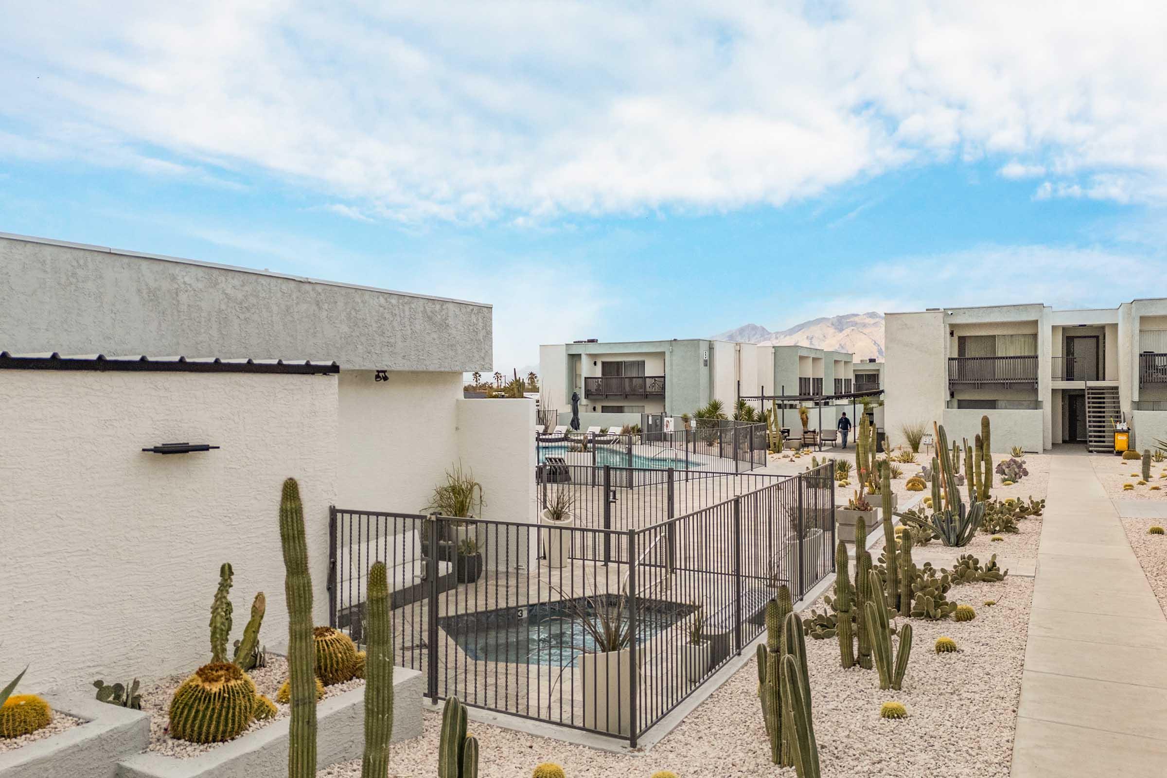 View of a modern desert resort featuring white buildings, a fenced pool area, and neatly arranged landscaping with cacti and gravel. The backdrop includes mountains under a clear blue sky, creating a tranquil and inviting atmosphere.