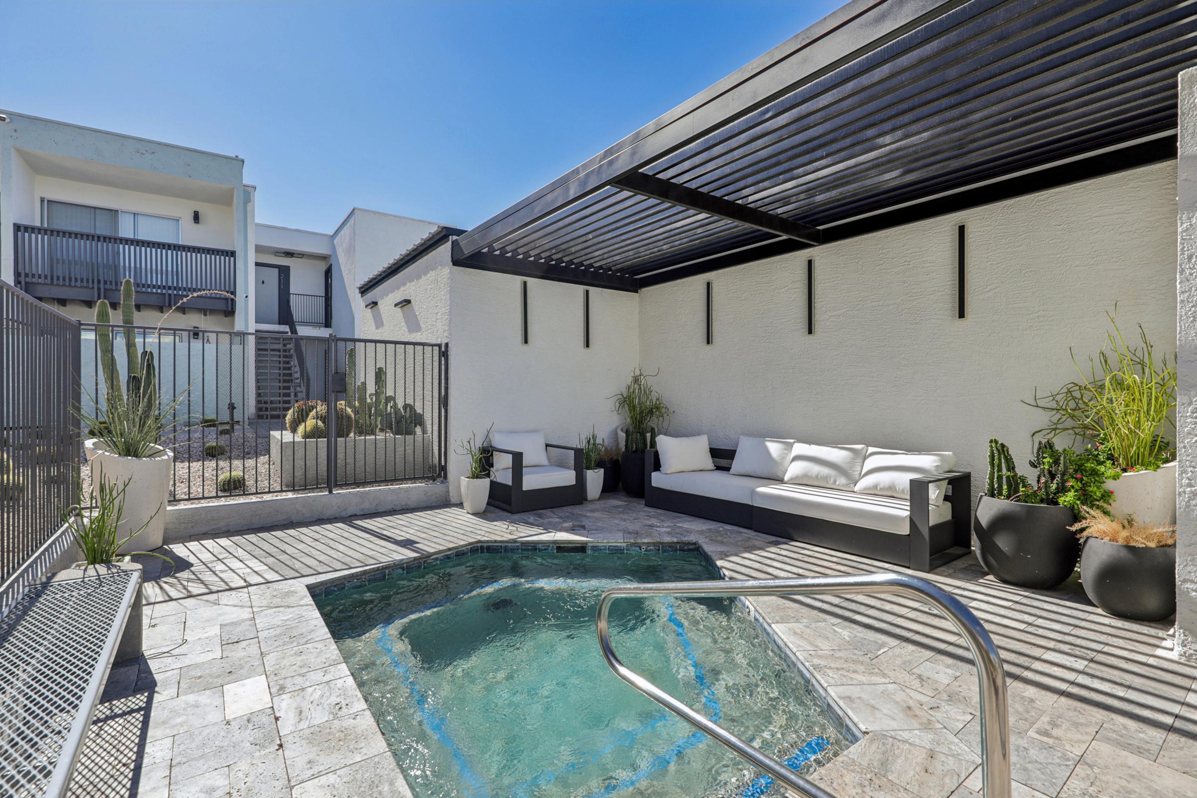 A modern outdoor space featuring a small jacuzzi surrounded by stone tiles, potted plants, and a shaded seating area with white couches. The area is enclosed by a metal fence and showcases a clear blue sky above, creating a relaxing ambiance in a private setting.