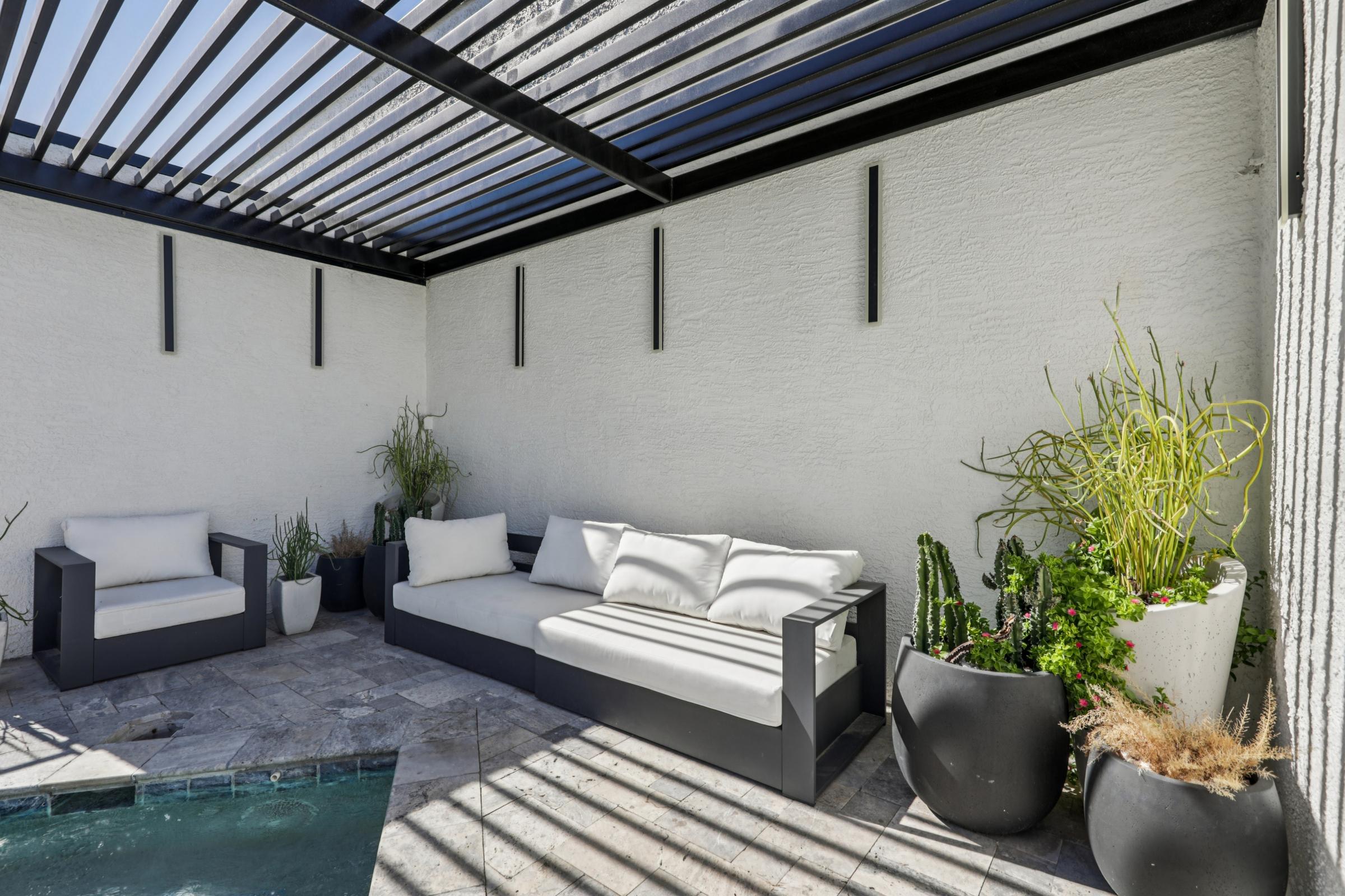 A modern outdoor lounge area featuring a shaded sectional sofa and an armchair, surrounded by potted plants. The space has a sleek stone floor and a small pool, with multiple vertical wall lights illuminating the white walls. The design emphasizes comfort and relaxation in a minimalist style.