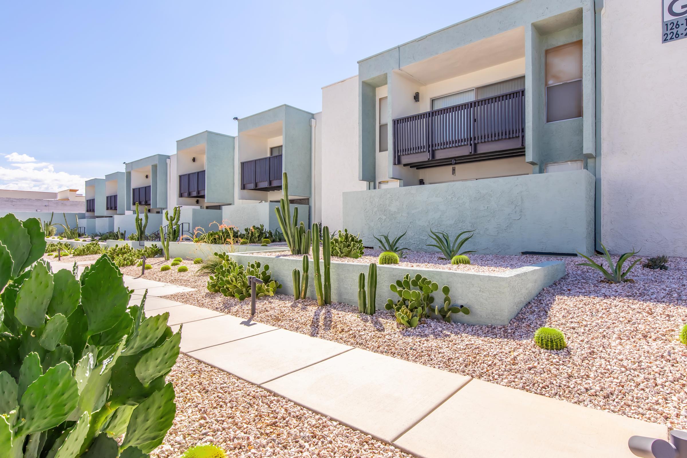 A modern building with three stories and balconies, set against a clear blue sky. The foreground features a landscaped area with various cacti and desert plants, surrounded by decorative gravel pathways leading to the entrances.
