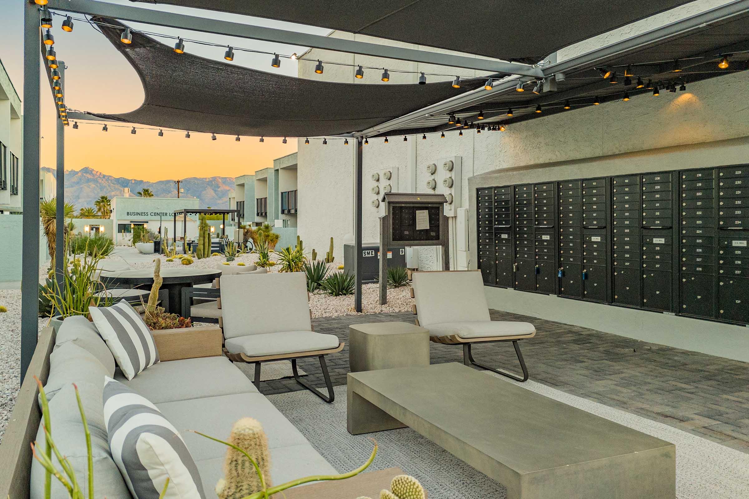 A modern outdoor lounge area featuring comfortable seating, a coffee table, and decorative cacti. In the background, there are mailboxes and a white building, with mountains visible under a colorful sunset. String lights are installed for ambiance.