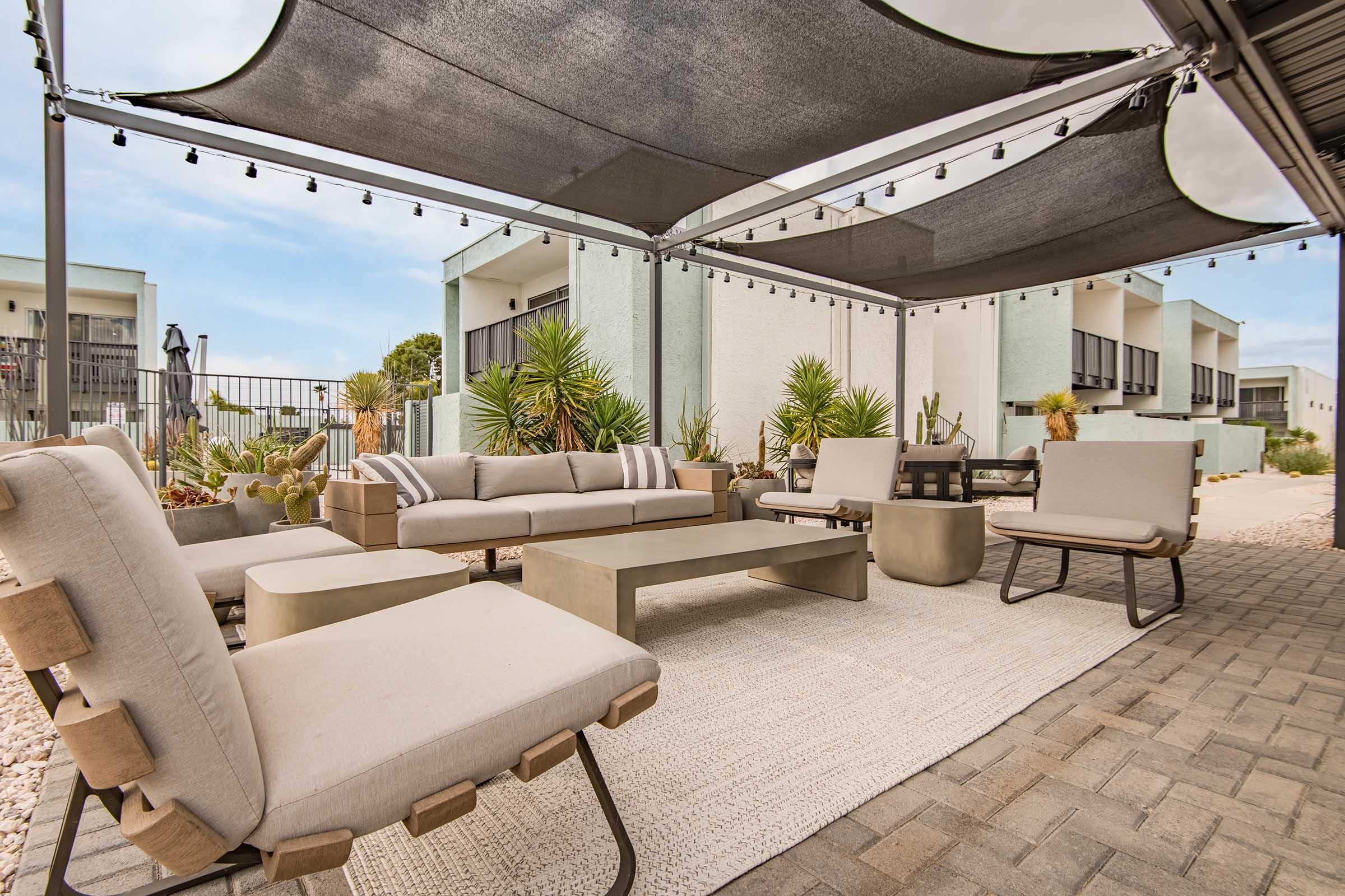 A modern outdoor lounge area featuring comfortable seating with neutral cushions, a low coffee table, and decorative plants. Shade sails provide protection from the sun, and string lights add ambiance. The background shows a minimalist building and landscaped space, creating a relaxing atmosphere.