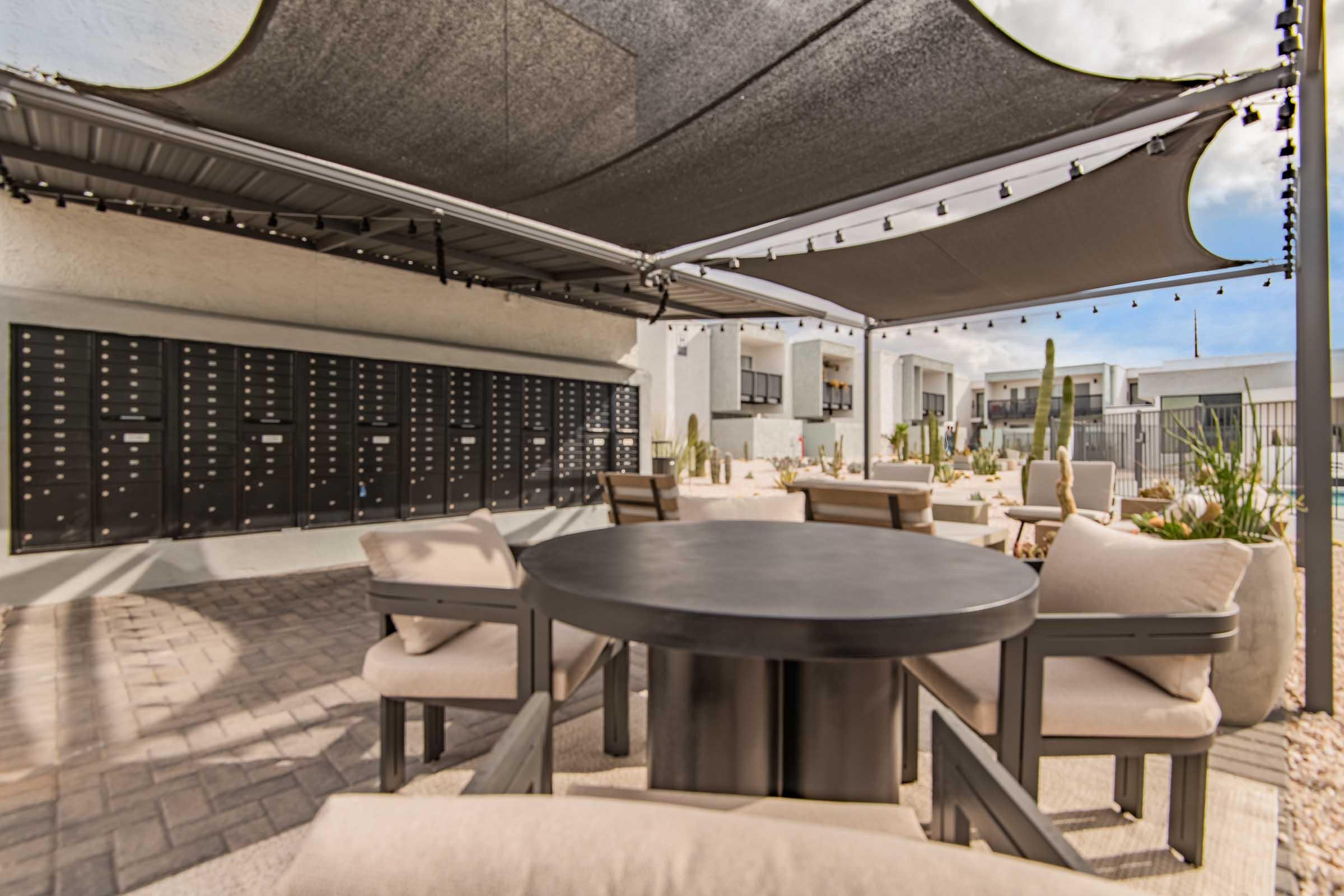 A shaded outdoor seating area features a round table surrounded by cushioned chairs. In the background, there are organized mailboxes against a wall, with desert landscaping, including cacti, adding to the ambiance. String lights are suspended overhead, creating a cozy atmosphere.