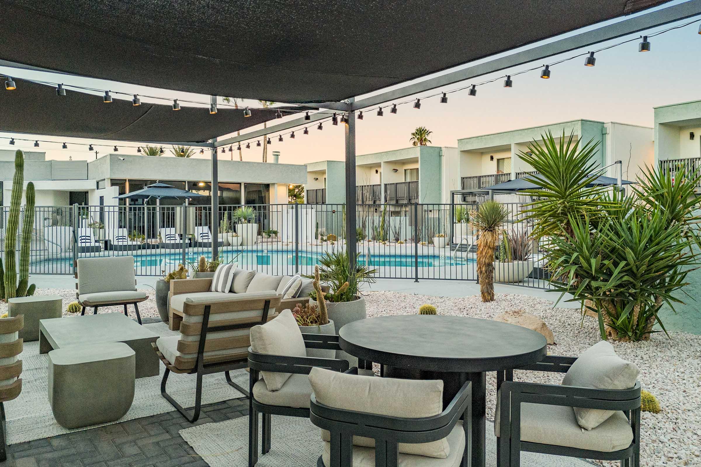 A modern outdoor lounge area featuring comfortable seating, a round dining table, and decorative cacti. The space is shaded by a canopy with string lights above, overlooking a swimming pool surrounded by sun loungers, with stylish buildings in the background. The atmosphere is inviting and tranquil.