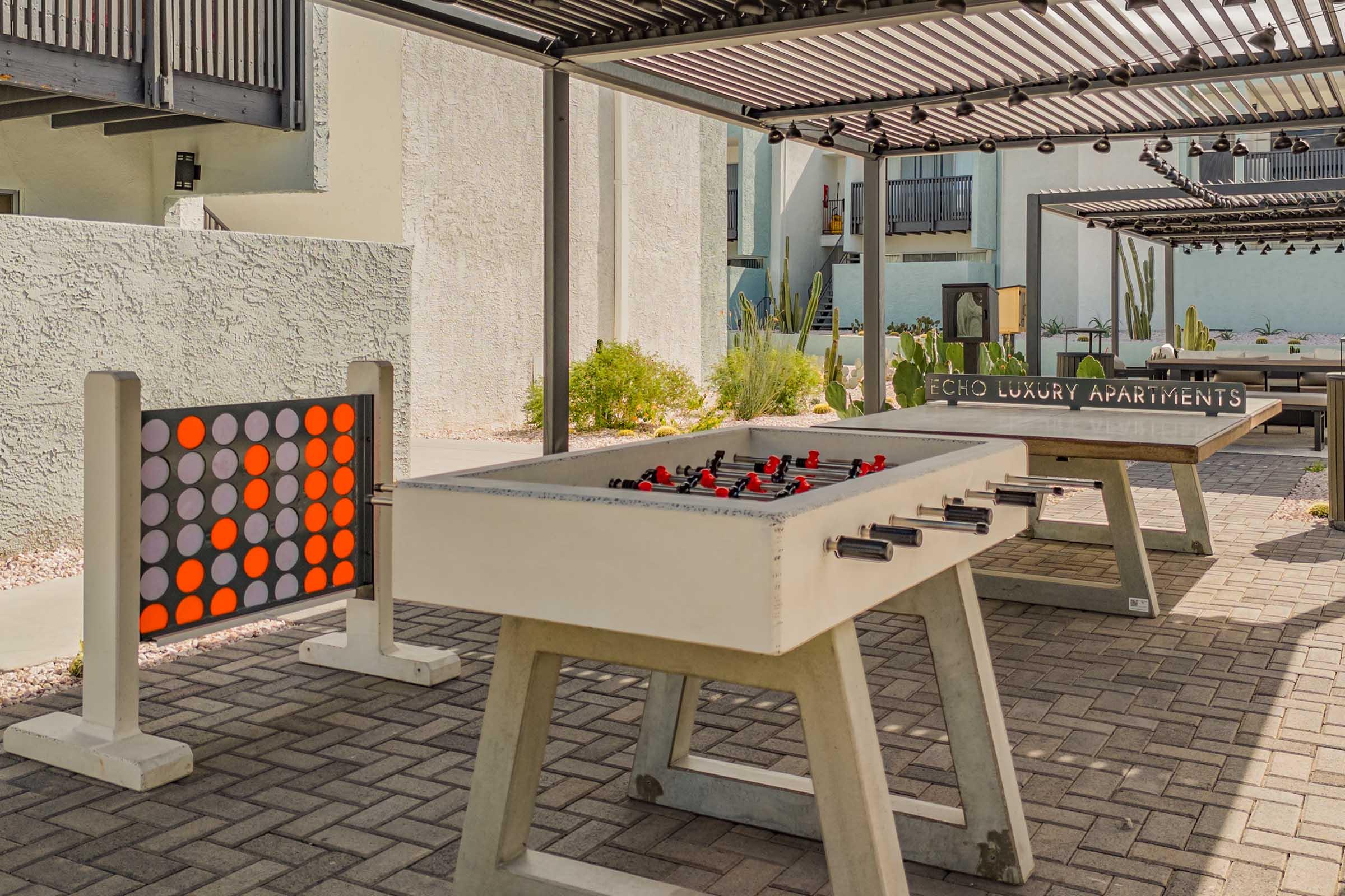 An outdoor recreation area featuring a foosball table and a colorful connect-four game. The background shows modern apartment buildings with shaded seating and landscaping, creating a vibrant community space at Echo Luxury Apartments.