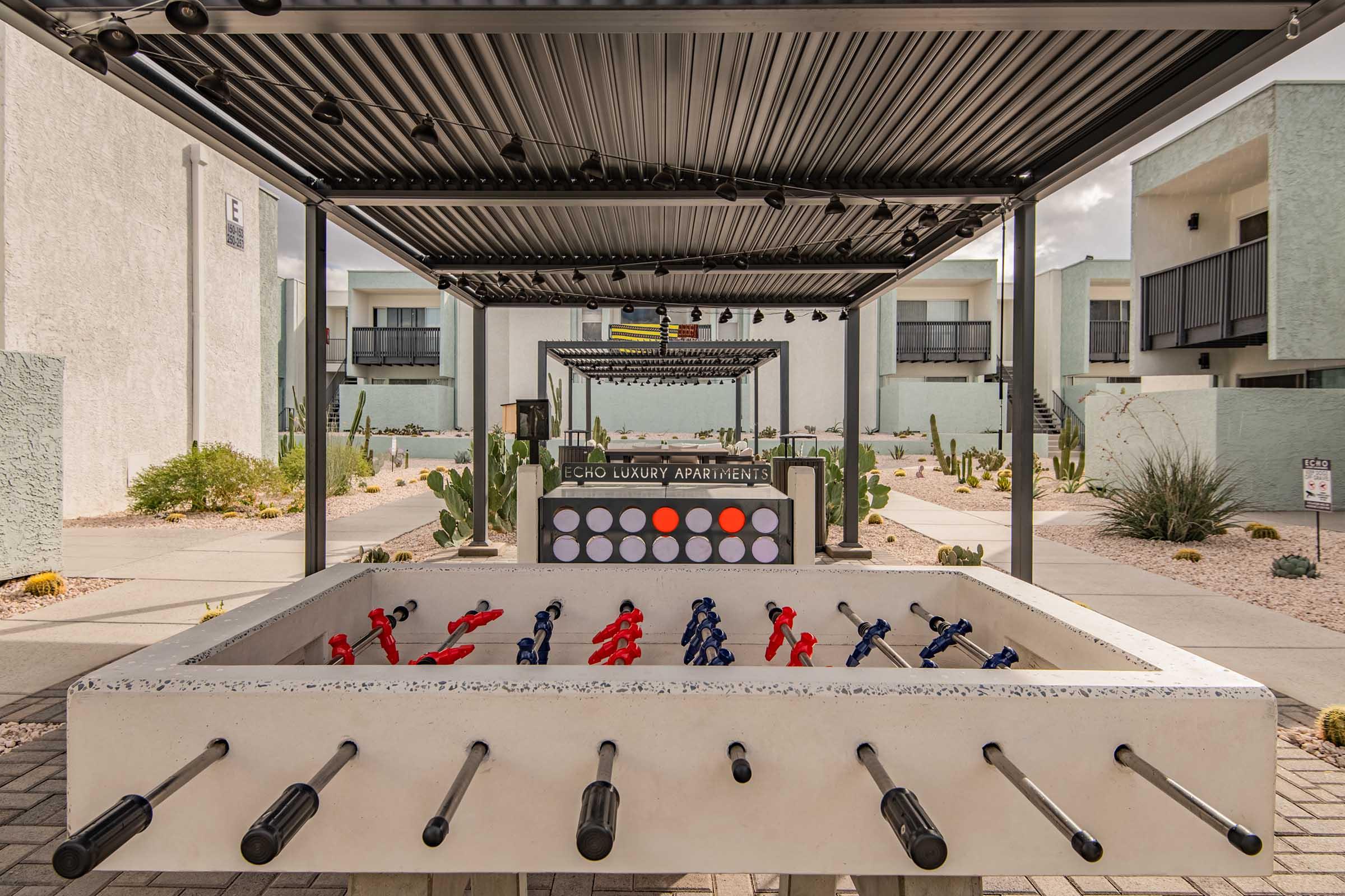 A large foosball table set outdoors in a landscaped area surrounded by modern apartment buildings. The space features cacti and a covered seating area, creating a recreational environment. Clear skies suggest a sunny day, enhancing the inviting atmosphere for residents and guests.