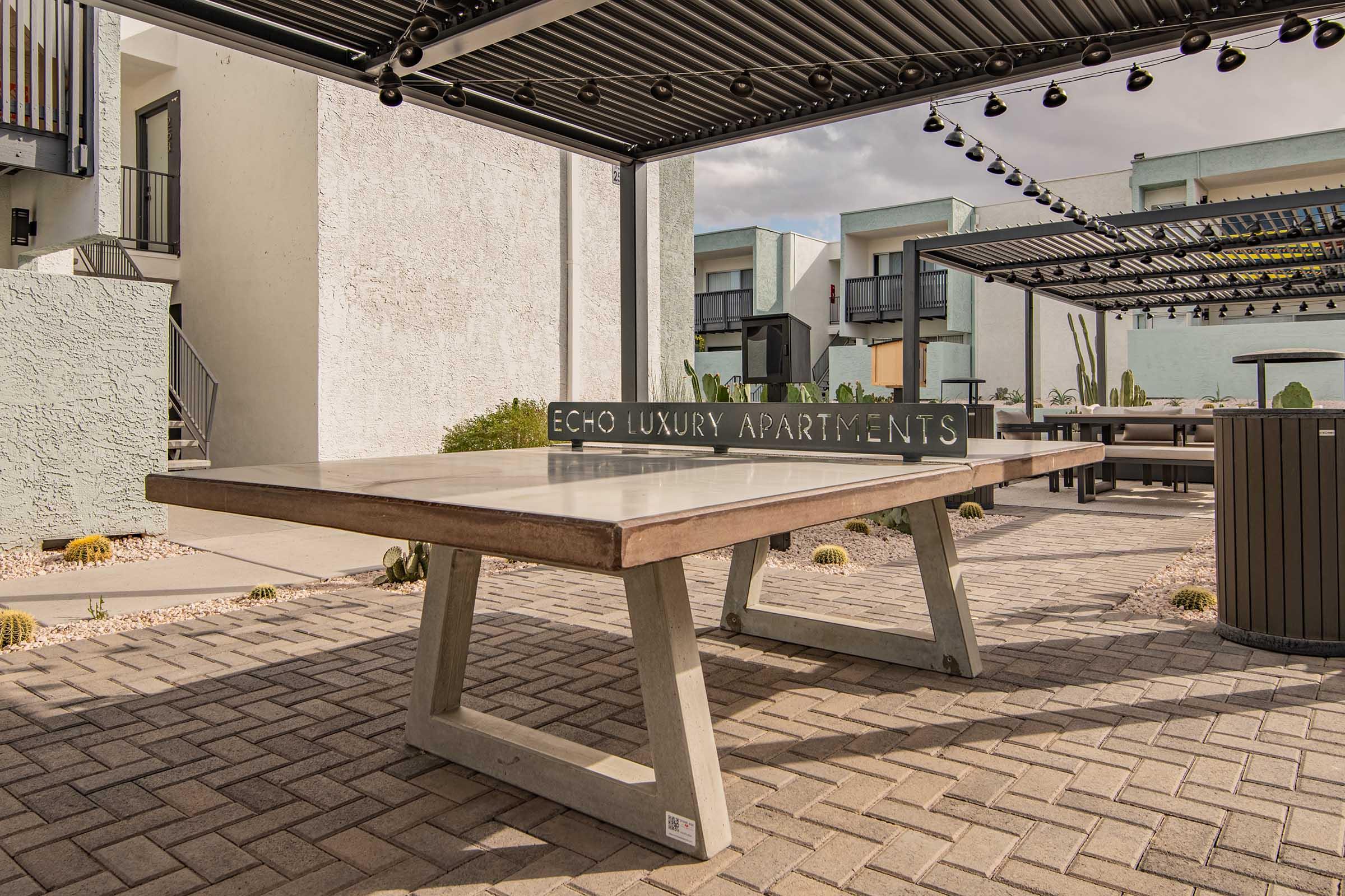 Outdoor seating area with a large rectangular table and a sign reading "ECHO LUXURY APARTMENTS" in a modern residential complex. The background features contemporary apartment buildings, and string lights overhead enhance the ambiance. Desert landscaping surrounds the area, providing a relaxed atmosphere.