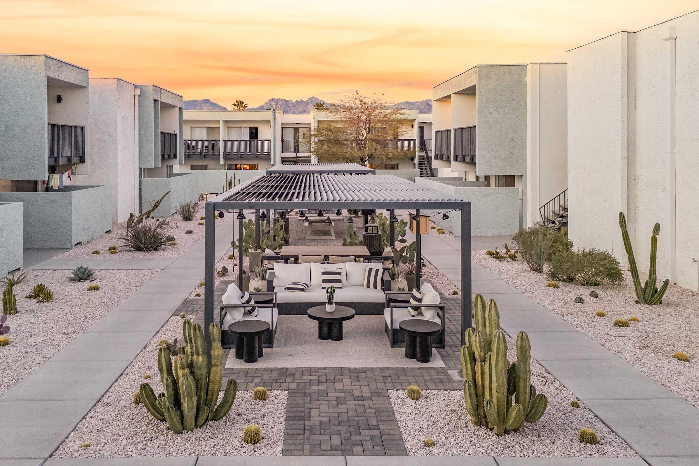 A modern outdoor lounge area featuring a pergola with comfortable seating, surrounded by desert landscaping with cacti and gravel paths. The backdrop includes minimalist architecture and a sunset sky, creating a tranquil ambiance in a residential complex.