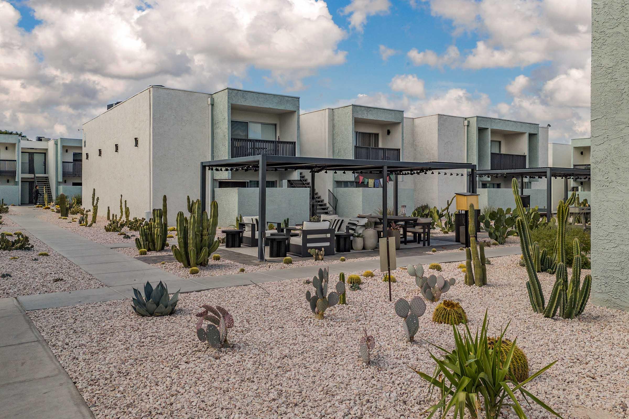 A modern apartment complex with white buildings and balconies, surrounded by landscaped desert gardens featuring various cacti and succulents. The pathway leads through the garden, with seating areas under shaded structures, all set against a backdrop of cloudy blue skies.