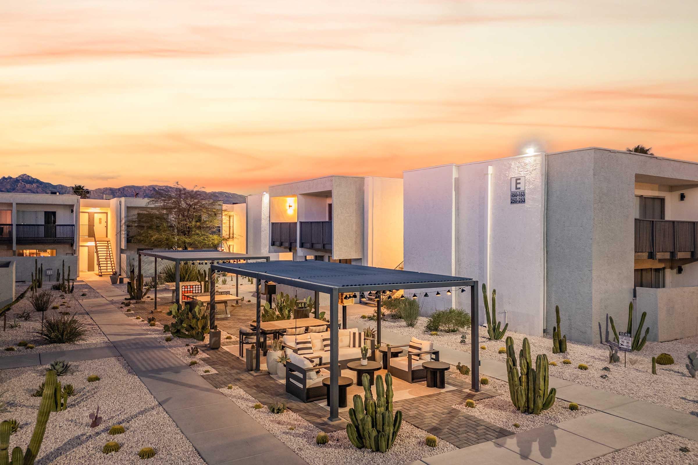 A desert-themed hotel exterior at sunset, featuring modern architecture with a tranquil courtyard. The area is adorned with desert plants and cacti, complemented by seating arrangements under shaded structures. The warm, colorful sky creates a serene atmosphere, highlighting the inviting charm of the setting.