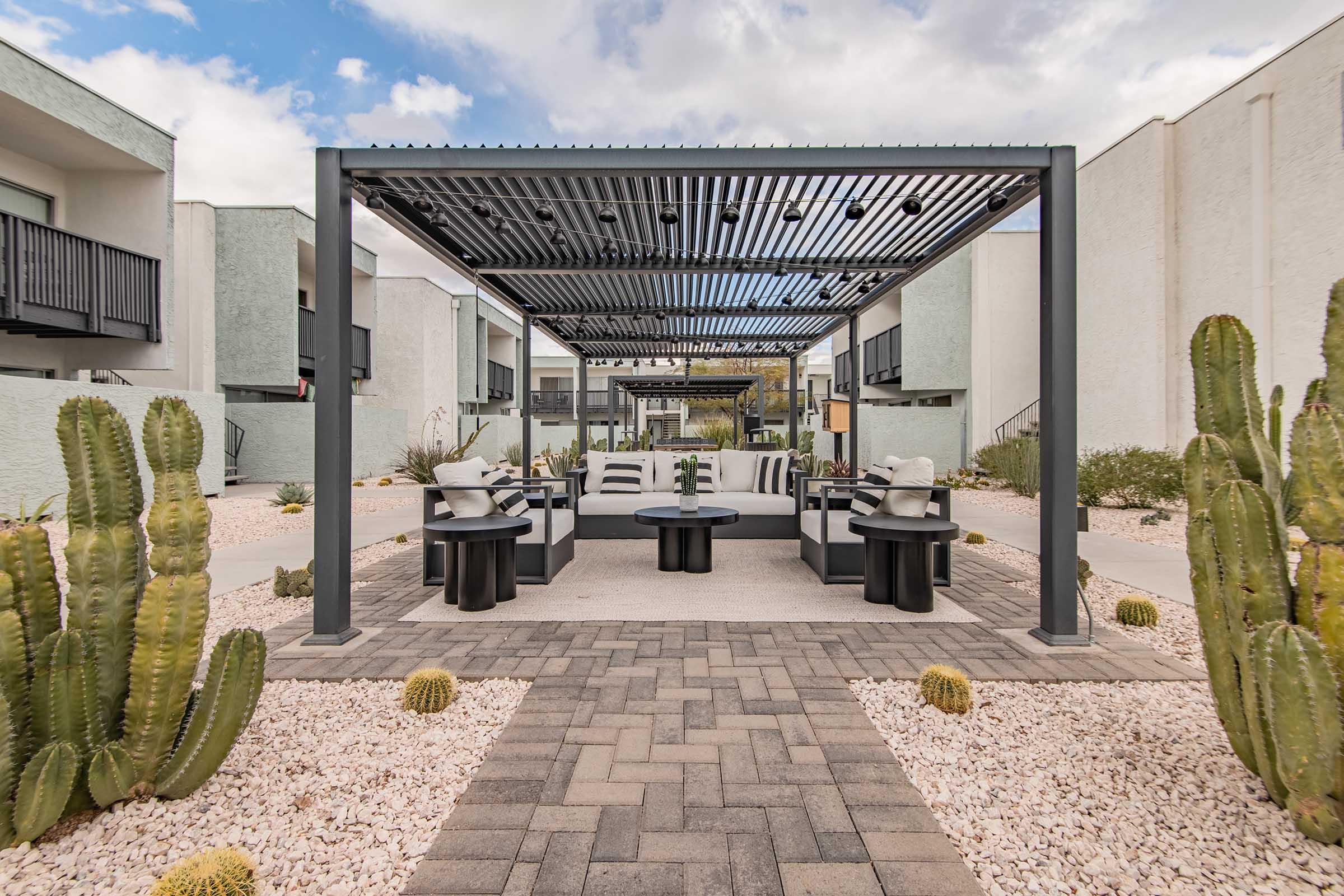 A modern outdoor seating area under a sleek pergola, featuring comfortable cushions on sofas and round tables. Surrounding the space are desert plants, including tall cacti and small decorative stones, set against a backdrop of contemporary buildings. The scene is bright and inviting, perfect for relaxation.