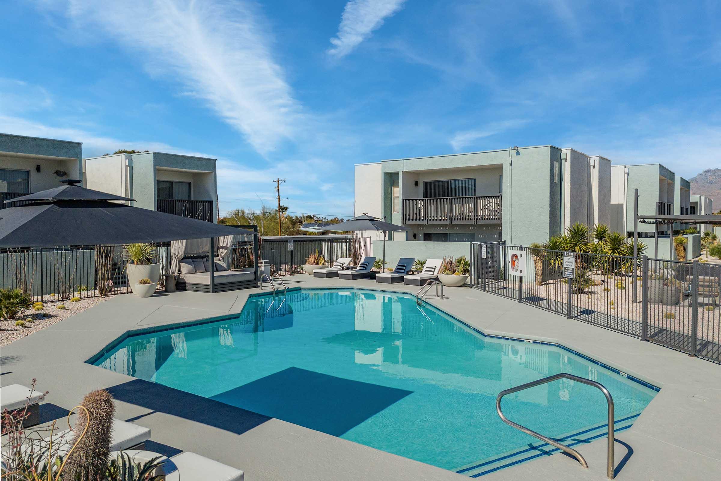 A clear blue swimming pool surrounded by lounge chairs, with a shaded seating area and modern buildings in the background. Palm trees and desert plants add greenery, while the sky features wispy clouds, creating a sunny, inviting atmosphere.