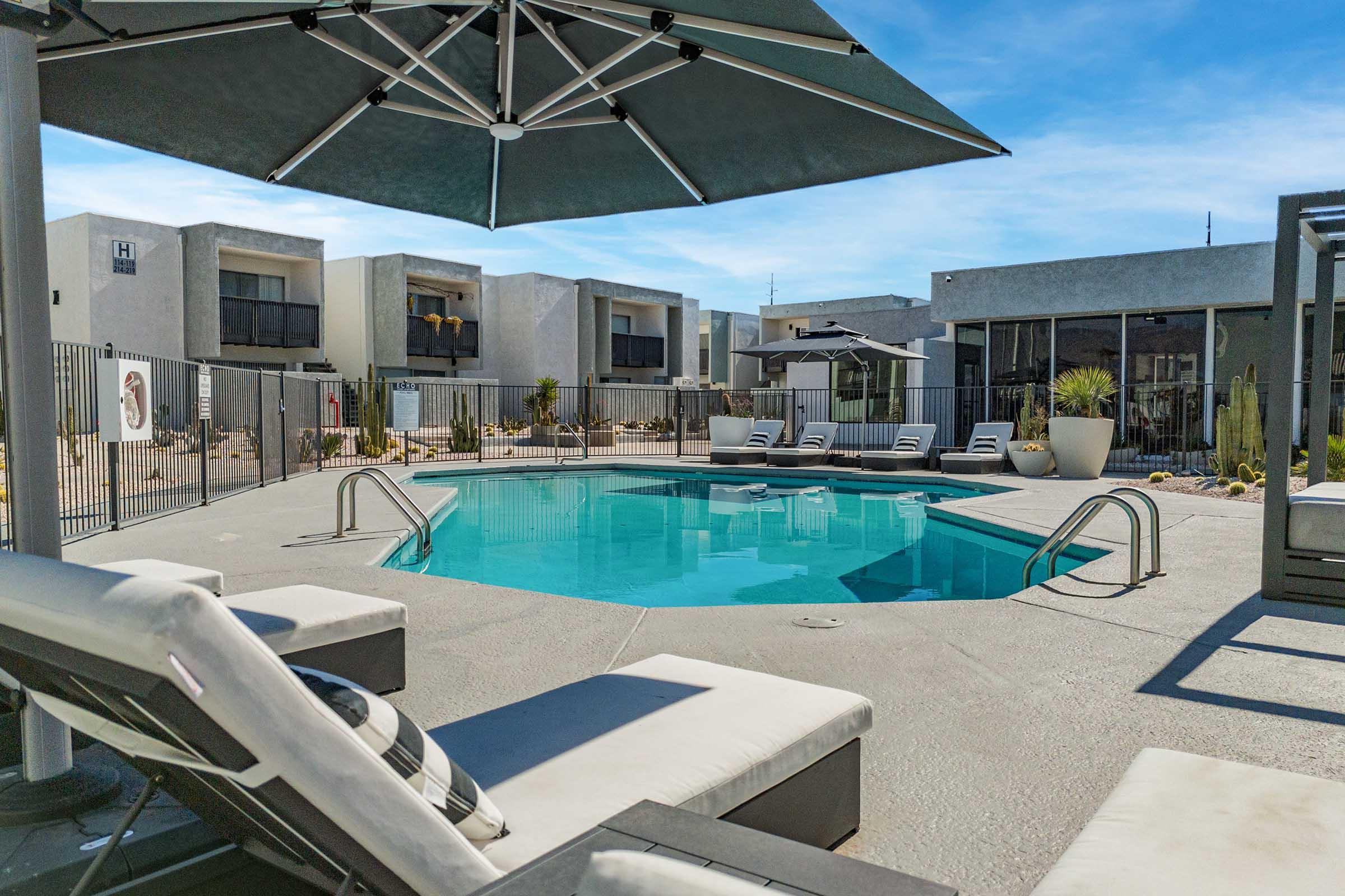 A modern outdoor pool area featuring a clear blue swimming pool surrounded by lounge chairs and a large umbrella. The setting includes simple, contemporary architecture in the background with a desert landscape, creating a relaxing atmosphere for leisure and enjoyment.