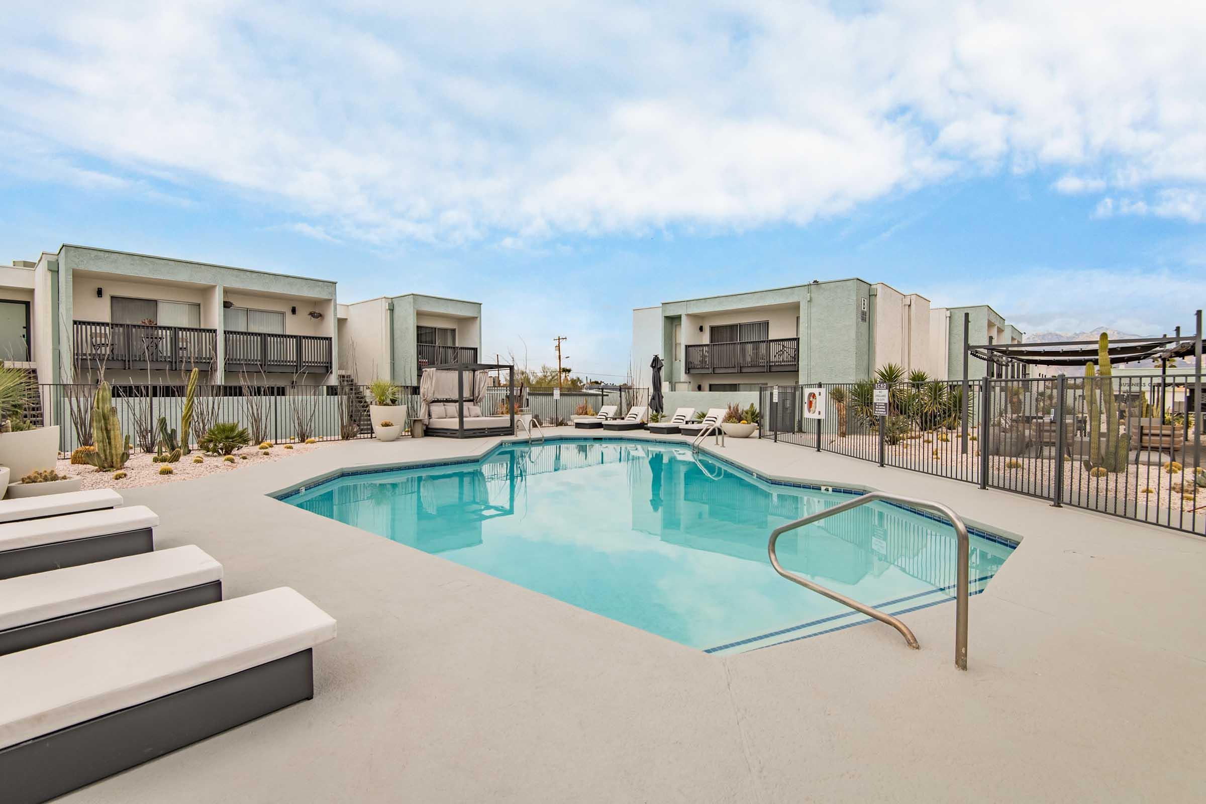 A modern outdoor pool surrounded by lounge chairs and desert landscaping. The pool is rectangular and features a staircase for entry. In the background, there are two-story buildings with balconies, and a clear blue sky above, creating a serene and inviting atmosphere.