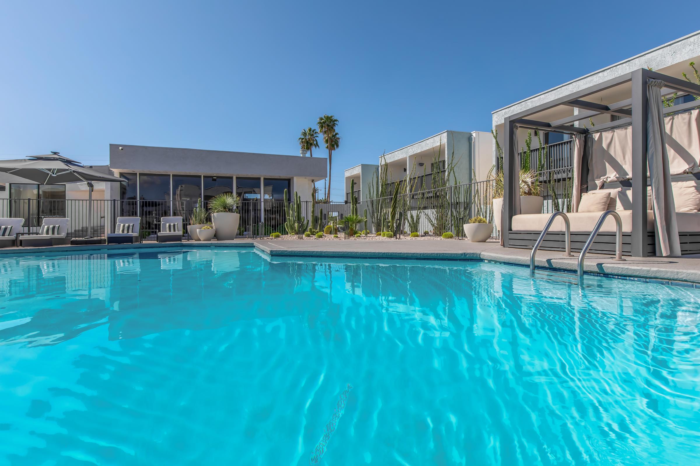 A clear blue swimming pool surrounded by lounge chairs and elegant cabanas, with palm trees nearby and modern buildings in the background under a bright blue sky. The scene is tranquil and inviting, perfect for relaxation.