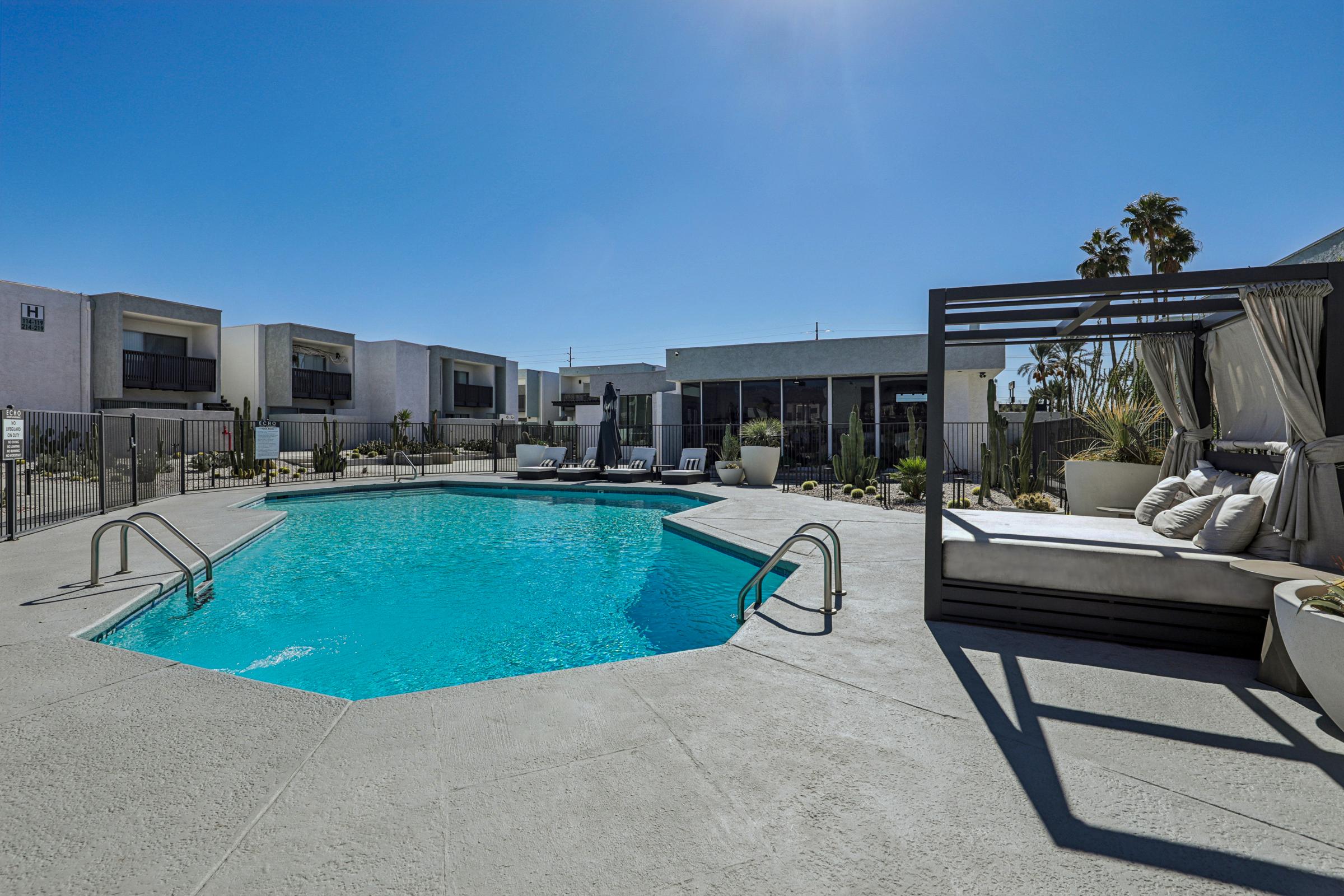 A bright outdoor pool area featuring a spacious pool with clear blue water. Surrounding the pool are lounge chairs and stylish cabanas, set against a backdrop of modern buildings and palm trees under a clear blue sky. The ambiance is inviting and perfect for relaxation.