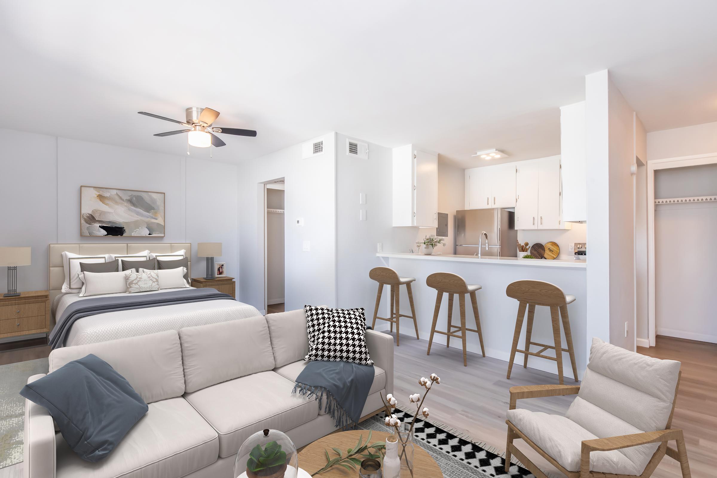 Modern living space featuring a light-colored sofa with throw pillows, a round coffee table, and stylish accent chairs. In the background, a neatly made bed with decorative pillows is visible, along with a kitchen area that includes a bar with stools. A ceiling fan and artwork add to the contemporary aesthetic.