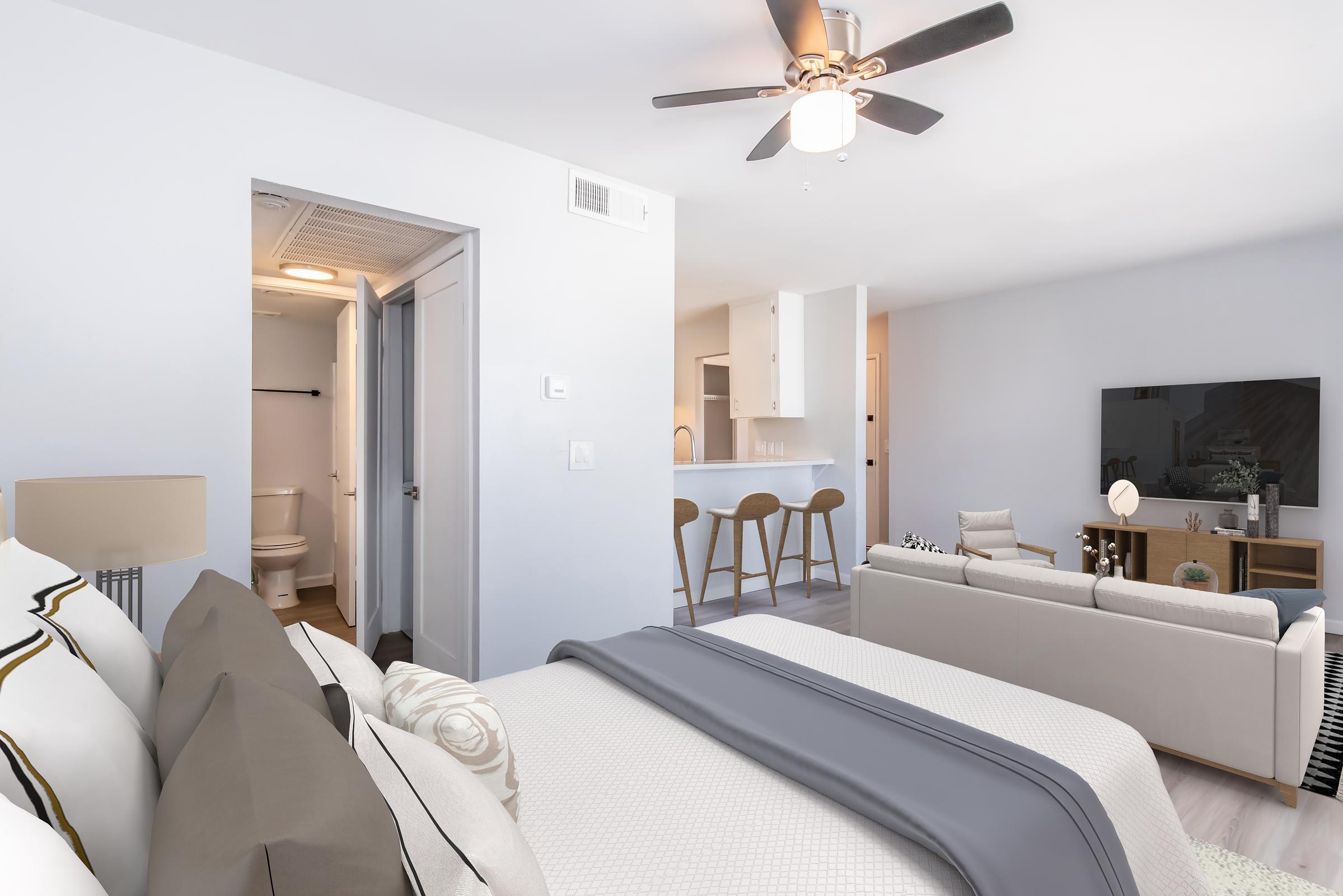 A modern, cozy living space featuring a neatly made bed with gray bedding, a stylish sofa, a ceiling fan, and a large TV on a media console. The room has light-colored walls, an open layout, and visible access to a bathroom, enhancing the inviting atmosphere.