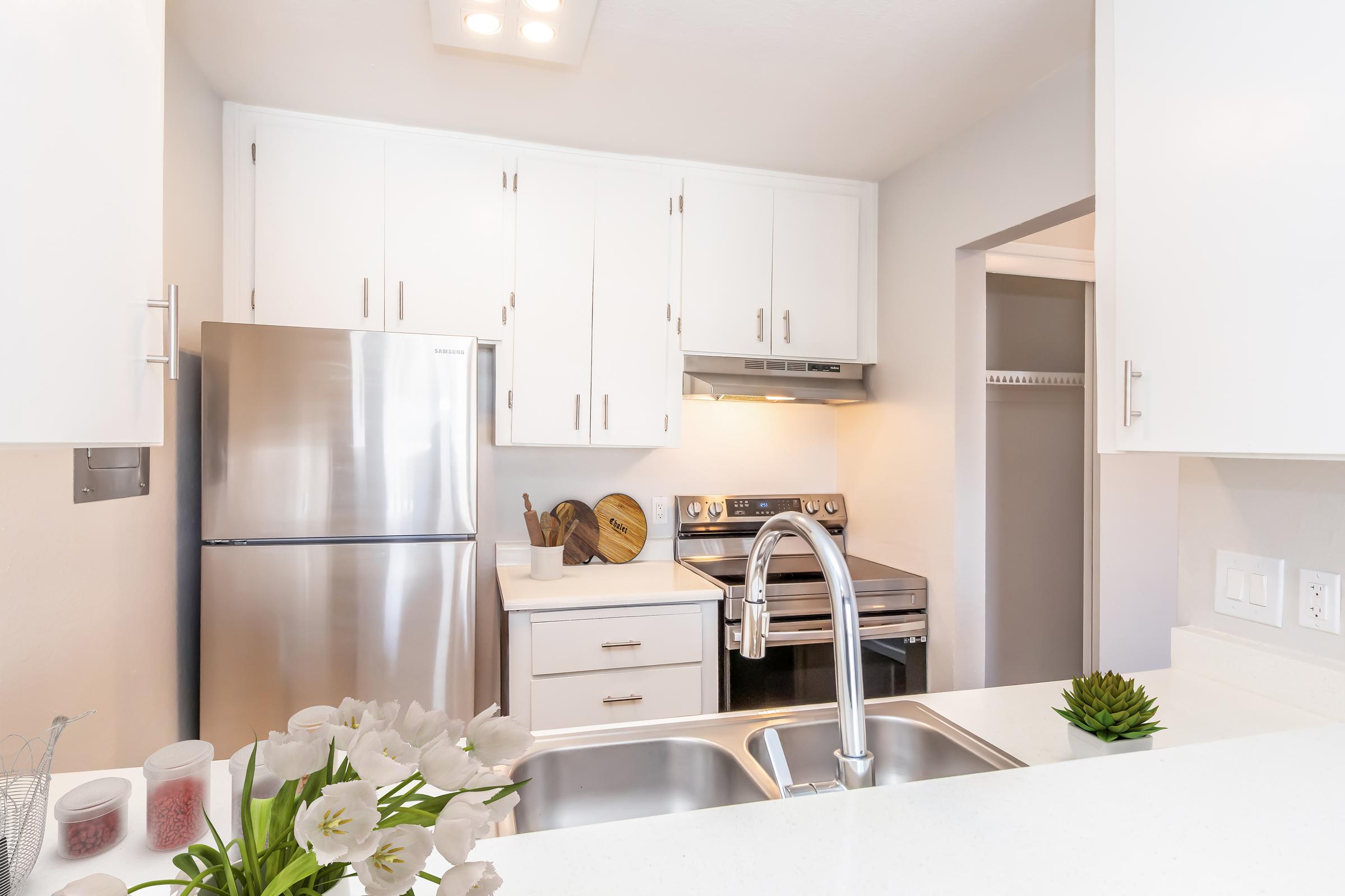 A modern kitchen featuring white cabinetry, stainless steel refrigerator, and an oven with a stove. A double sink is visible in the foreground, accompanied by a small plant and a vase of flowers. The kitchen appears bright and well-organized, with light-colored countertops and a clean aesthetic.