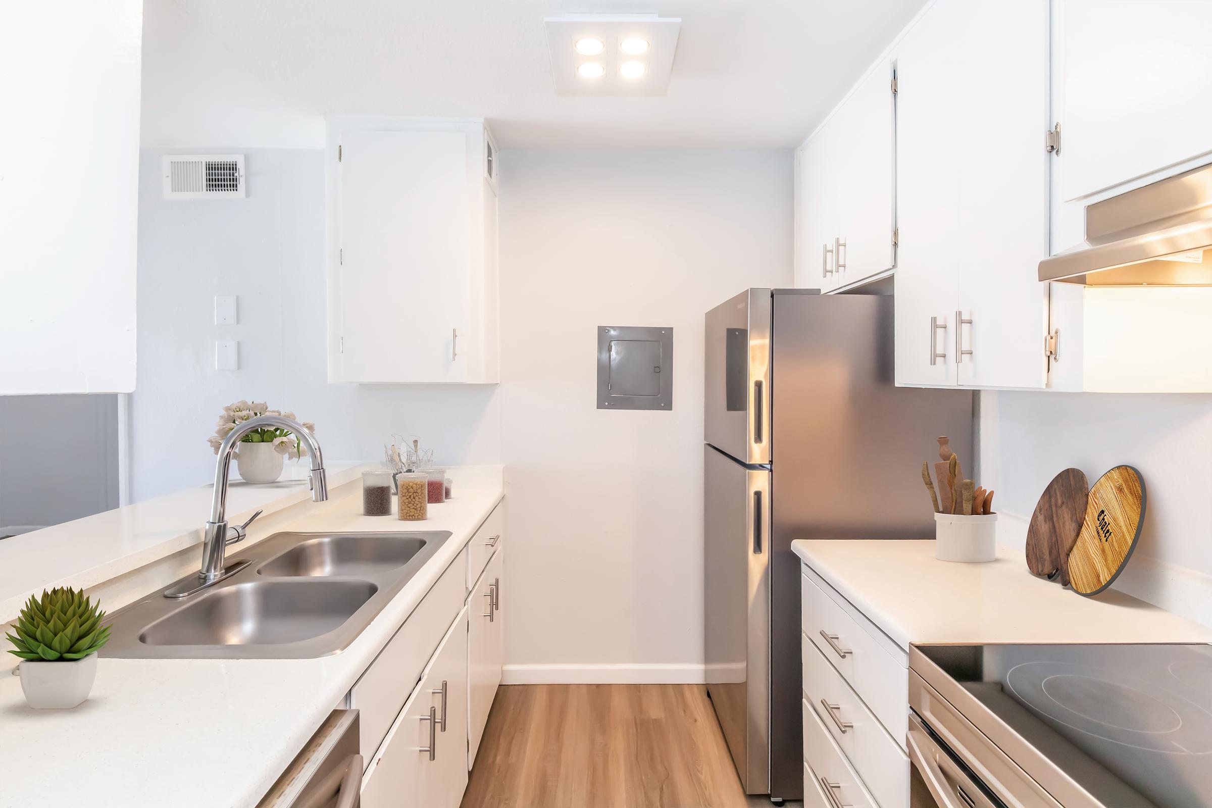Modern kitchen featuring white cabinetry, stainless steel appliances including a refrigerator, a double sink, and a countertop with cooking utensils and a small plant for decoration. The flooring is light wood, and the space is well-lit with recessed lighting above.