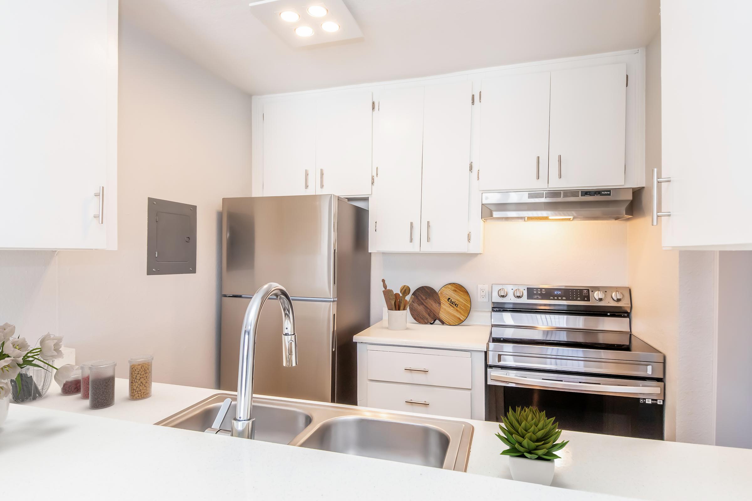 Modern kitchen featuring white cabinetry, stainless steel appliances including a refrigerator and stove, a double basin sink, and a small succulent plant. Wooden cutting boards are displayed on the countertop, along with containers of grains and a simple decorative touch. The space is bright and well-organized.