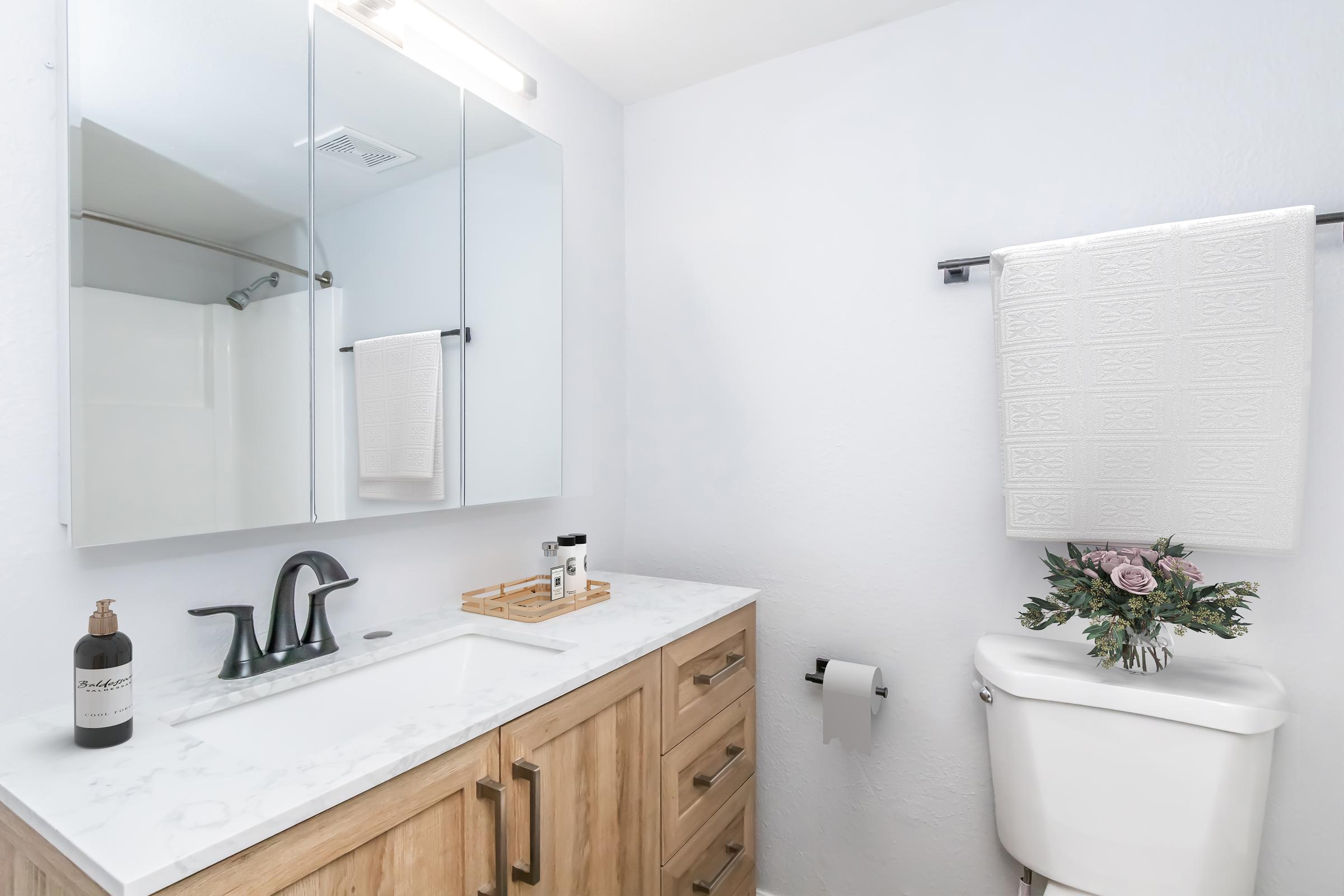 A modern bathroom featuring a double vanity with marble countertops, a large mirror, and stylish black fixtures. On the counter, there are toiletries and a wooden tray. A white towel hangs on a rod, and a decorative bouquet in a vase is placed next to the toilet. The walls are painted light and bright.