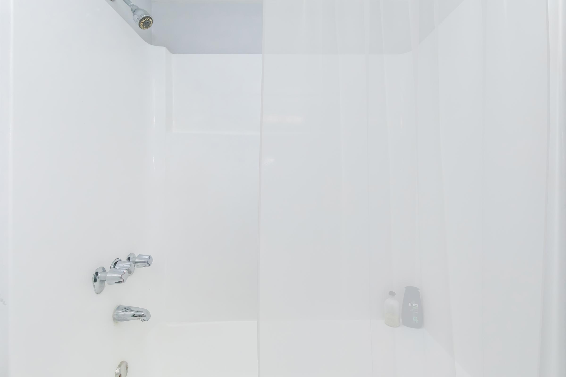 A clean, modern shower space featuring white walls and a transparent shower curtain. The water controls are visible, along with two bottles of shower products on the floor. The overall ambiance is bright and minimalistic.