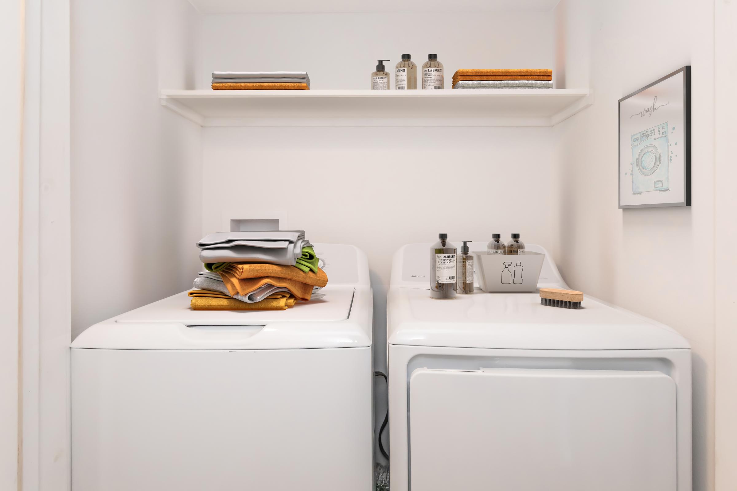 A clean laundry area featuring a washing machine and a dryer side by side. On top of the washing machine, there are neatly folded towels and a few laundry supplies. Above, a shelf holds decorative bottles and a framed picture on the wall adds a touch of style to the space.