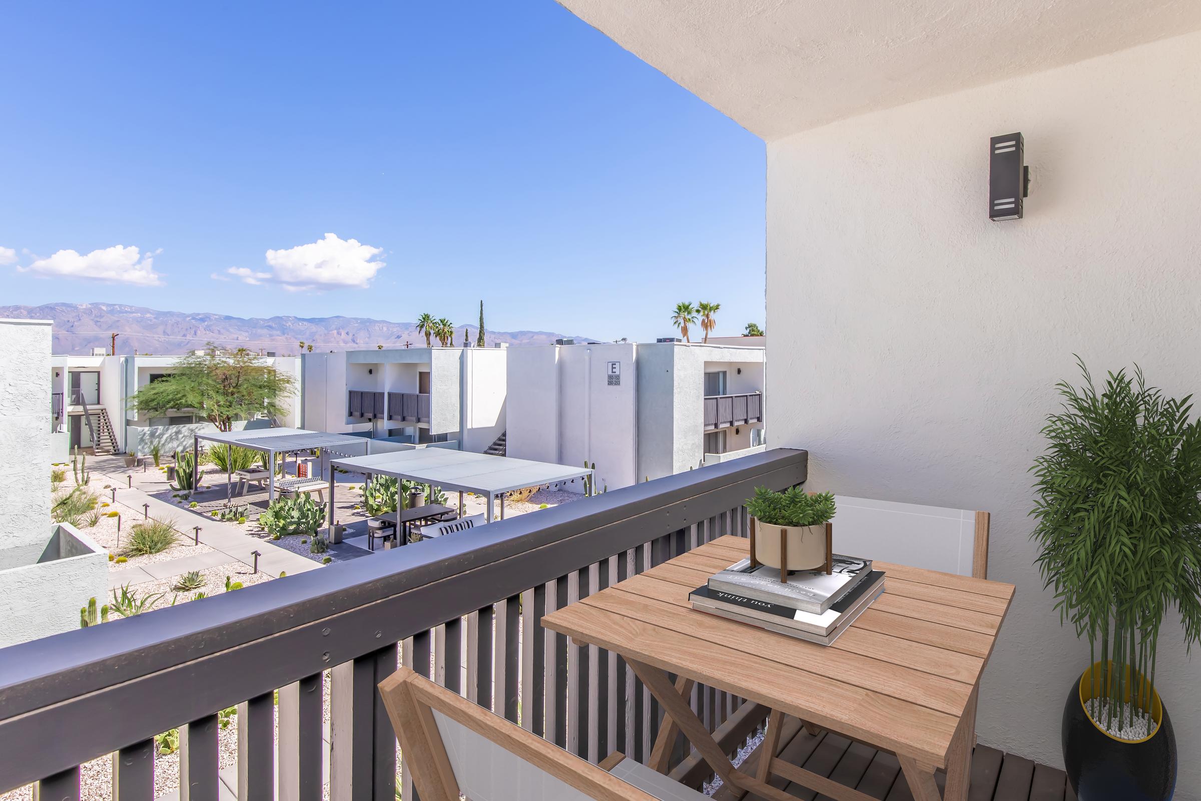 A balcony view featuring a wooden table with potted plants, surrounded by modern architecture and palm trees. In the background, mountains and a clear blue sky with a few clouds are visible, creating a peaceful outdoor atmosphere.