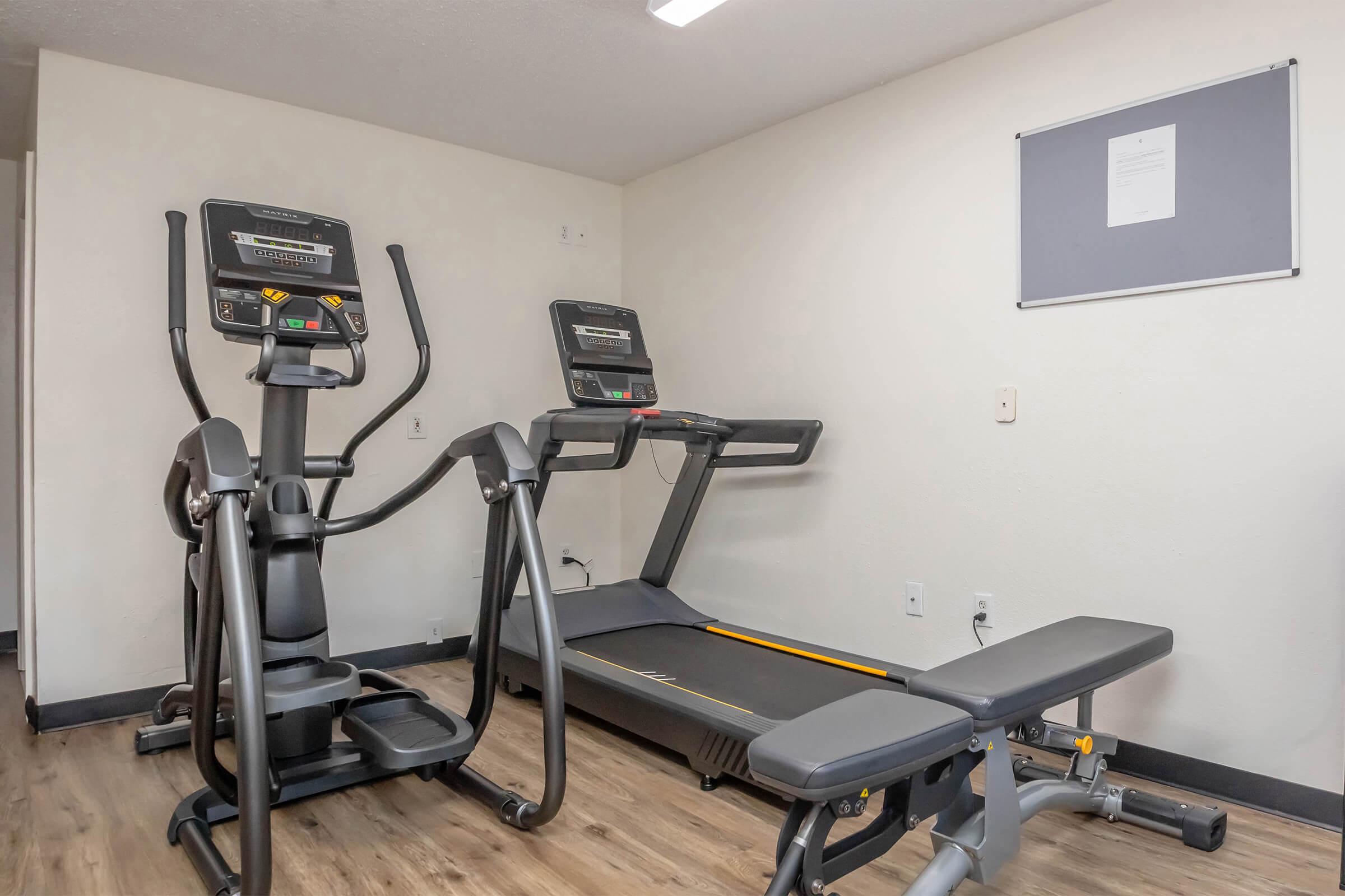 A fitness room containing a treadmill, an elliptical machine, and a weight bench. The equipment is set against a light-colored wall, with a gray bulletin board above. The flooring is wooden, and the room appears bright and well-lit.