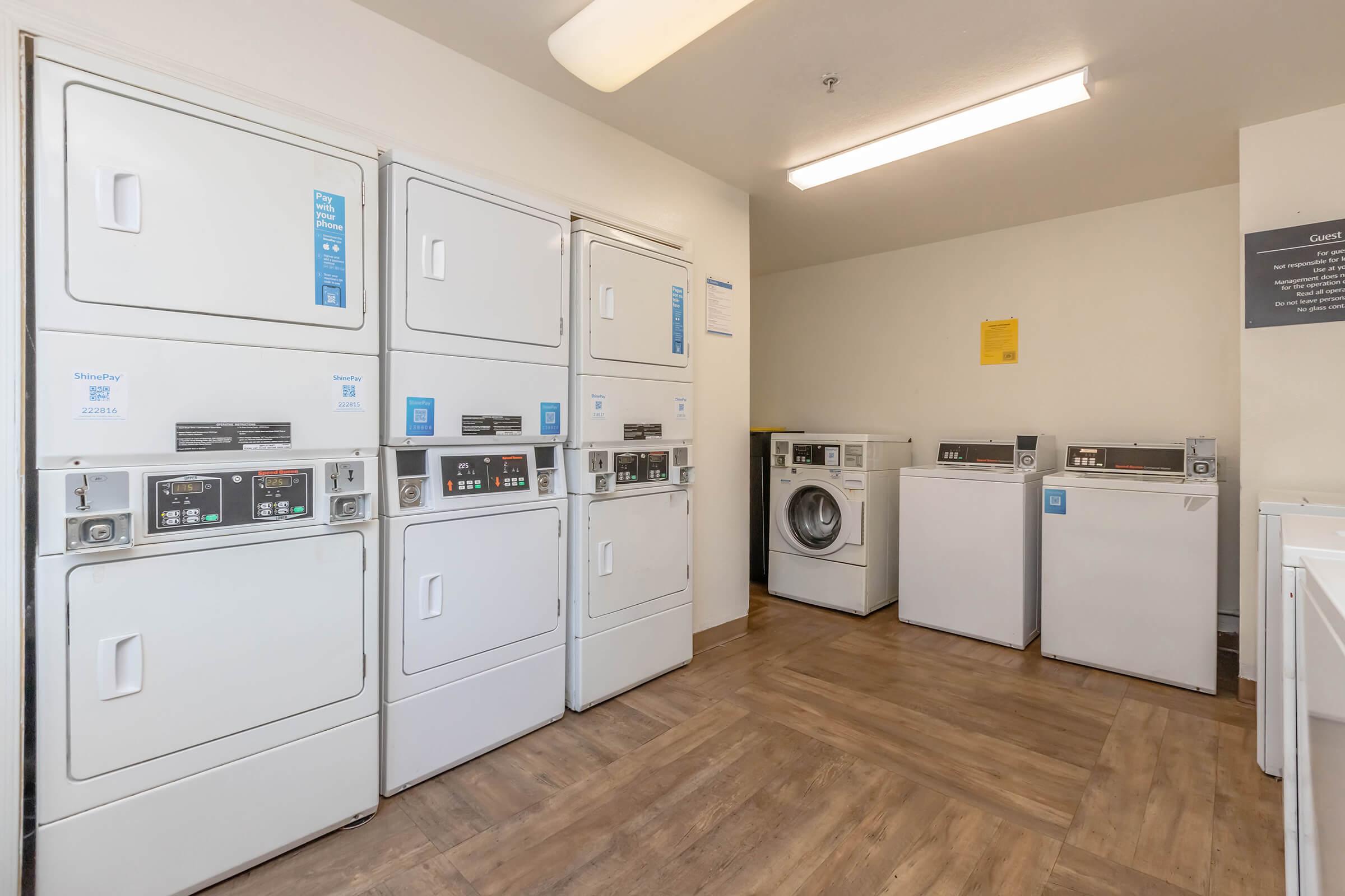 A laundry room featuring multiple stacked washer and dryer units along one wall, along with a front-loading washing machine and dryer on the opposite side. The floor is wood-like, and there are signs on the walls providing instructions. Bright, even lighting fills the space.