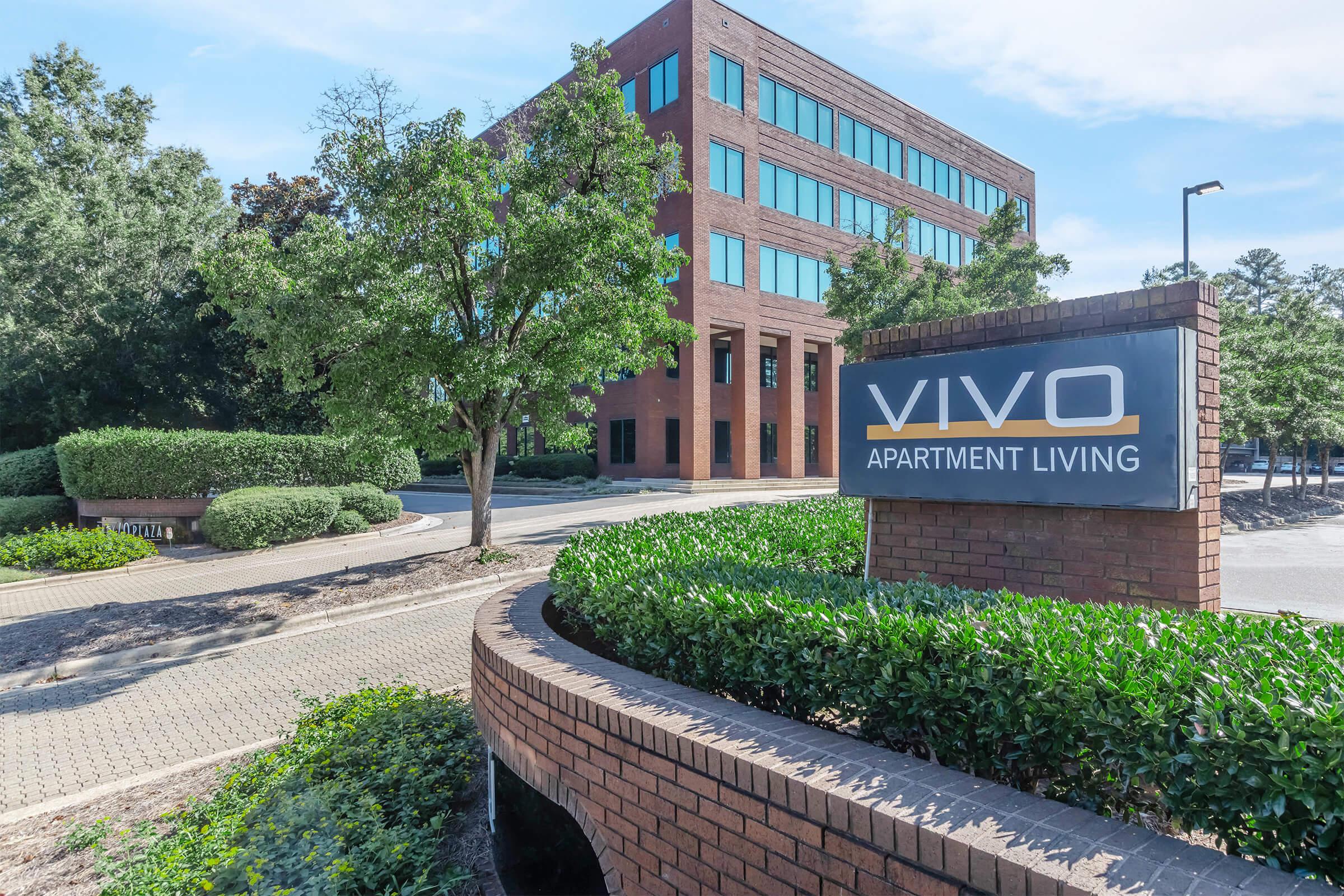 Sign for Vivo Apartment Living located in front of a modern brick building, surrounded by lush greenery and landscaping, with a clear blue sky above. The area features well-maintained pathways and shrubs, suggesting a welcoming residential environment.