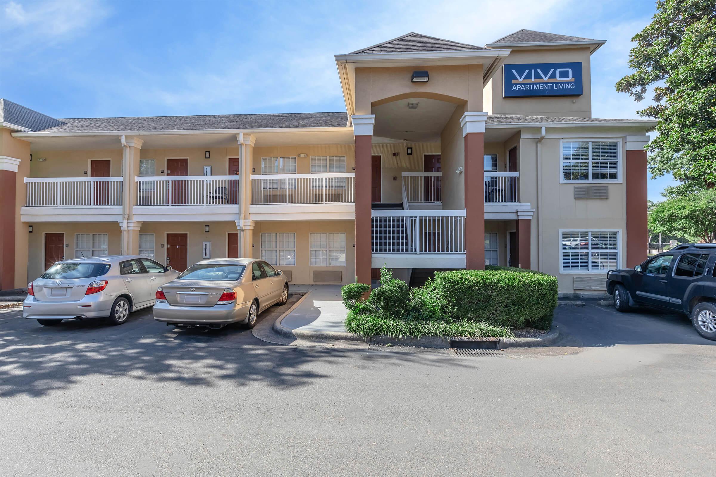 Front view of a two-story apartment building named "VIVO Apartment Living." The structure features multiple balconies with white railings and is flanked by parked cars in the foreground. Lush green shrubs line the base of the building, and bright blue sky fills the background, creating a welcoming atmosphere.