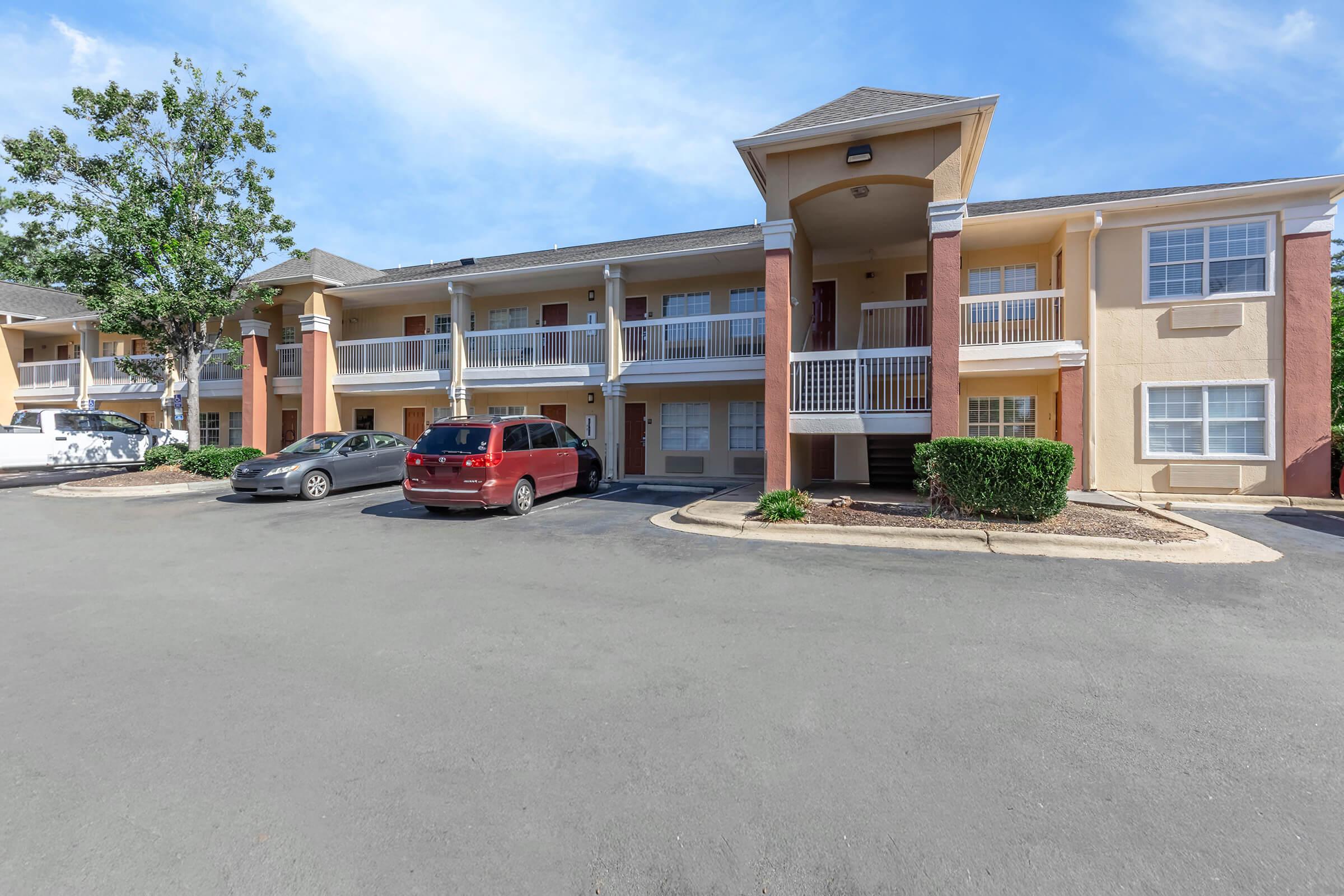 Exterior view of a two-story apartment building featuring multiple balconies. The parking lot in front has several cars parked. The building is painted in light colors with decorative pillars, surrounded by landscaped areas and trees under a clear blue sky.